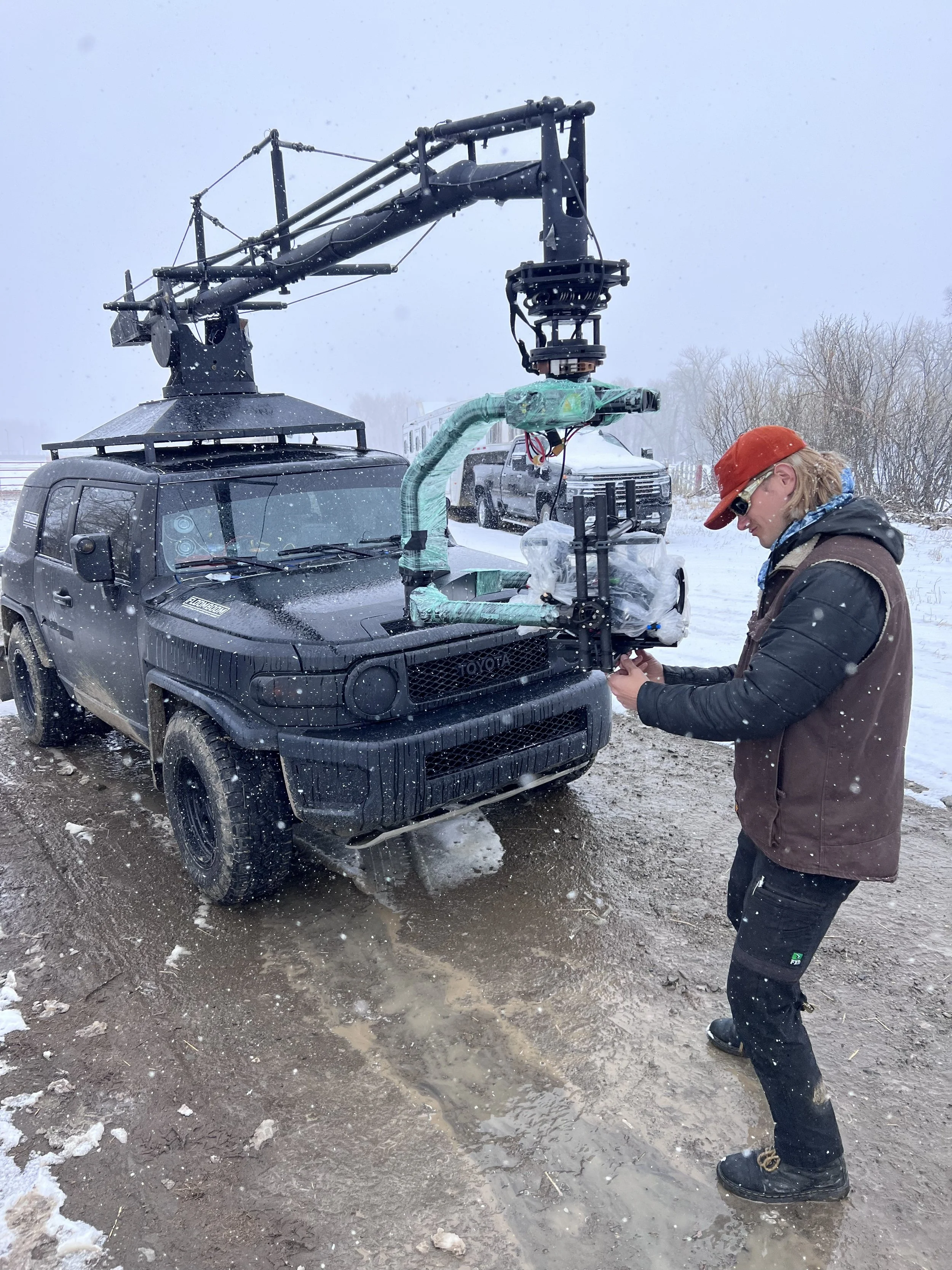 A person operating a camera mounted on a tripod attached to the front of a black off-road vehicle in snowy weather.