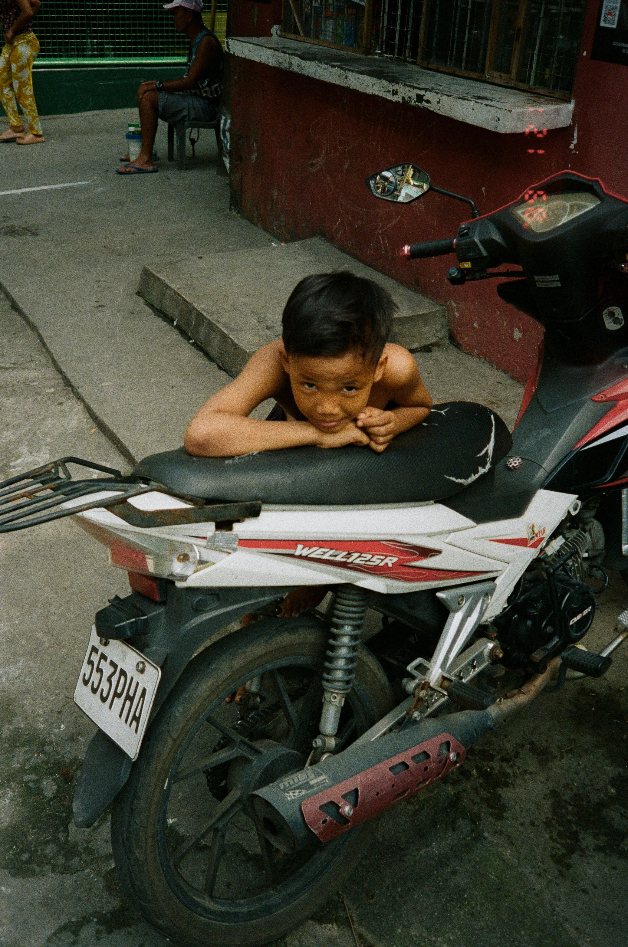 Young boy resting on a motorcycle parked on the sidewalk, with a red wall and a small window behind him, and a person sitting on a chair in the background.
