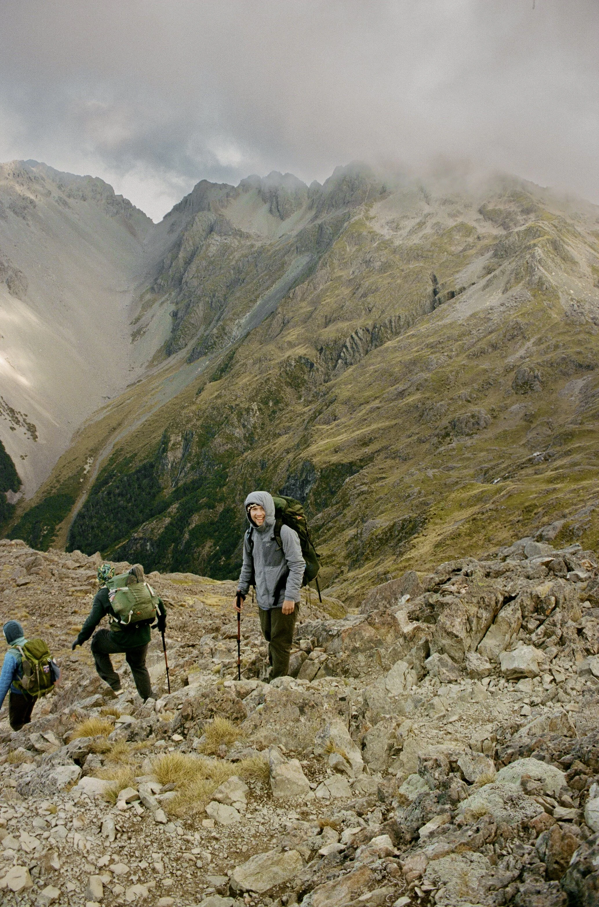 Group of hikers ascending a rocky mountain trail with steep slopes and mountain peaks in the background, some with clouds and fog.