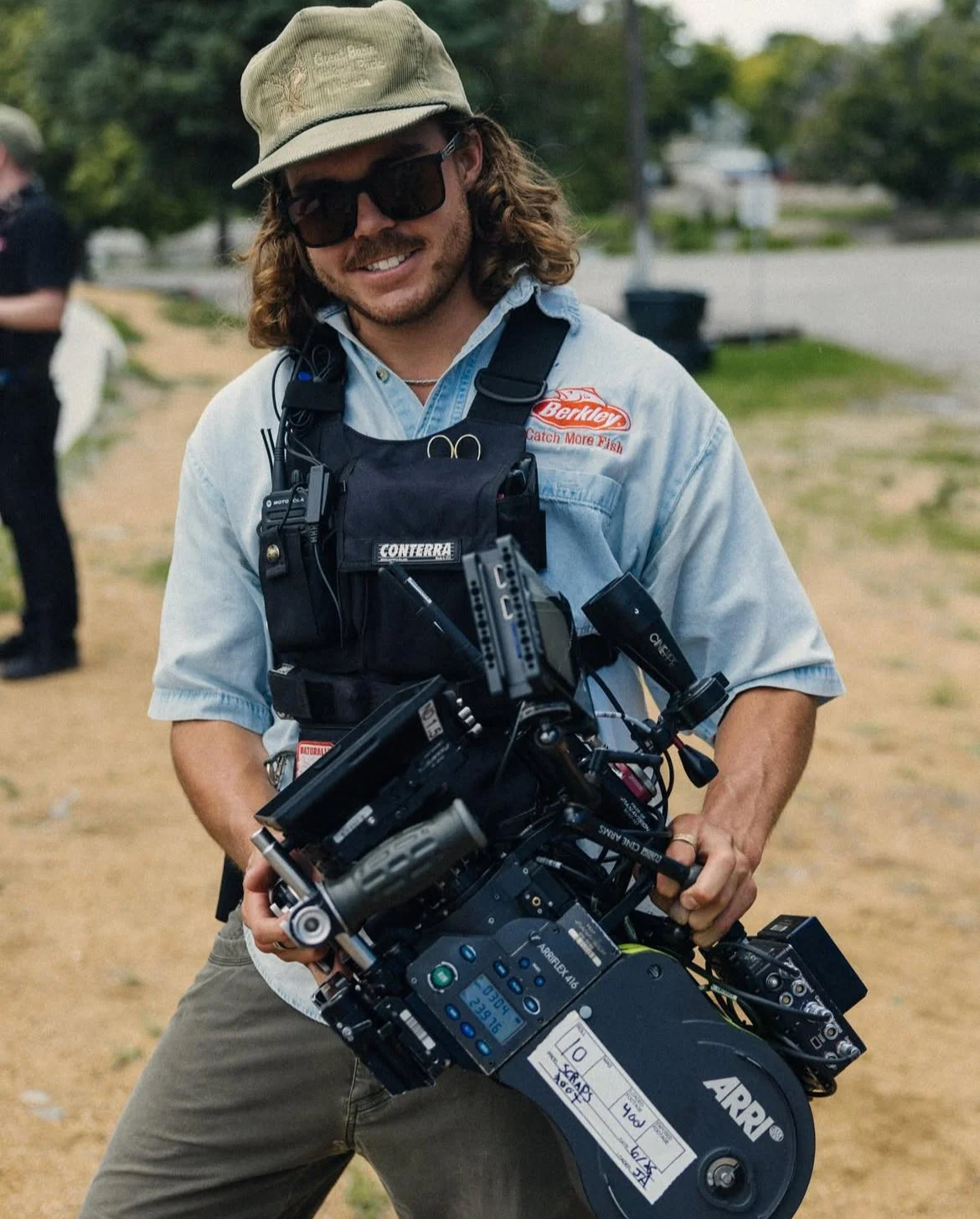 Man in sunglasses and hat holding a professional film camera with a smile, standing outdoors on a dirt field.
