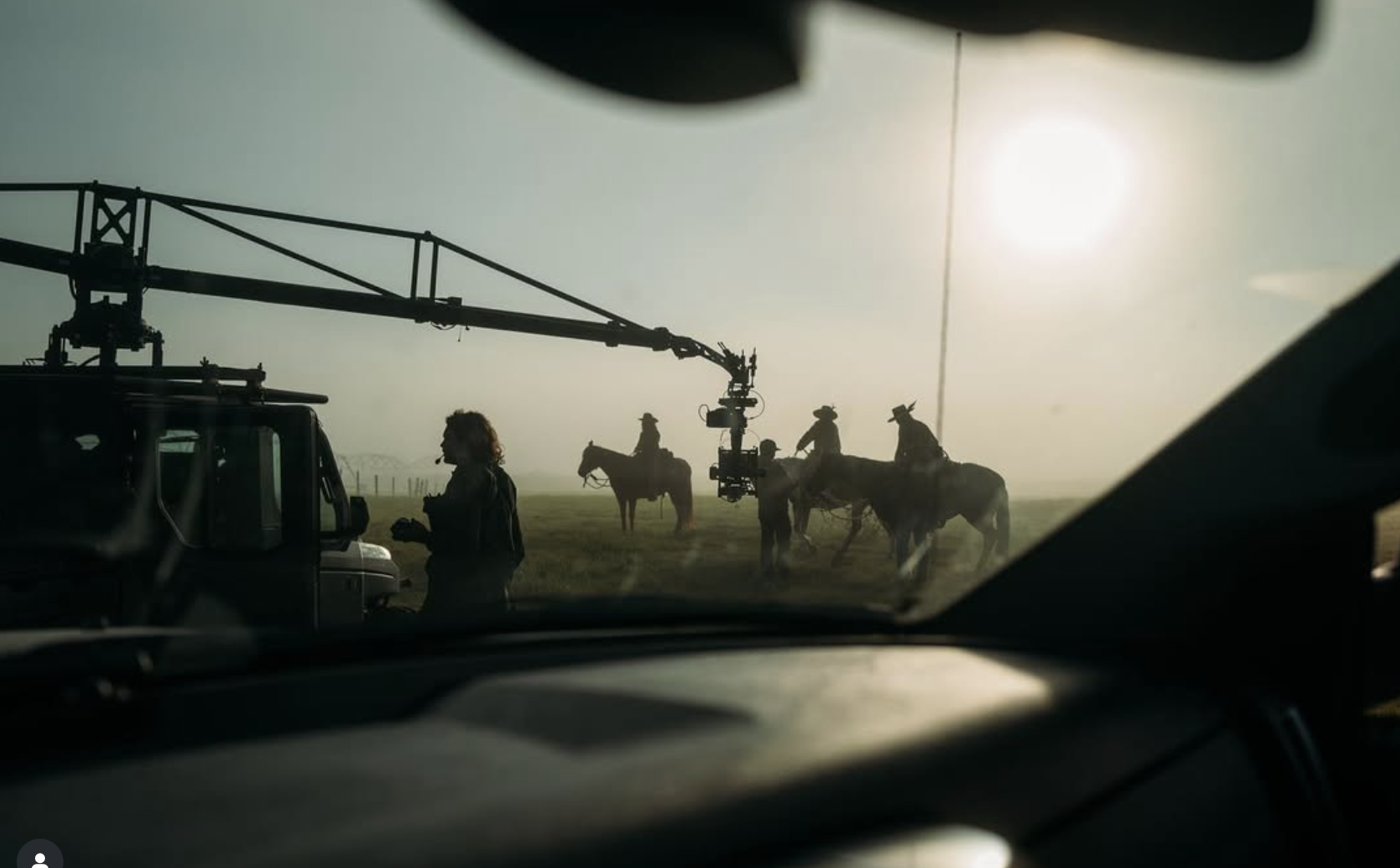 Silhouette of people on horseback and a person standing on the ground, seen through the windshield of a vehicle with a camera crane and a grassy field with a fence in the background, during sunrise or sunset.