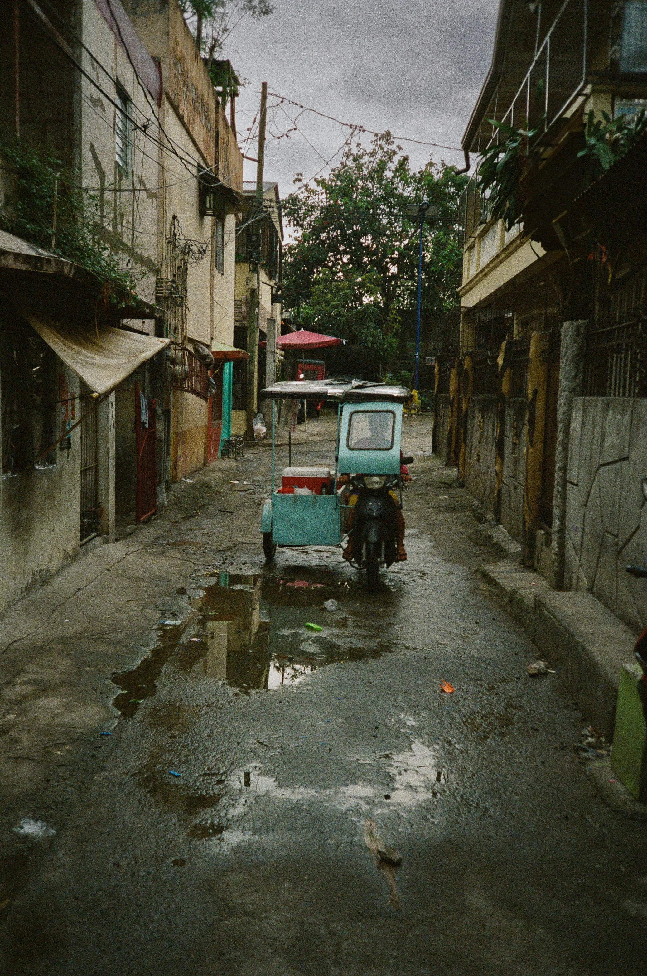 A narrow alleyway with a water puddle in the foreground, a small three-wheeled vehicle with a canopy in the middle, and old buildings on both sides under an overcast sky.