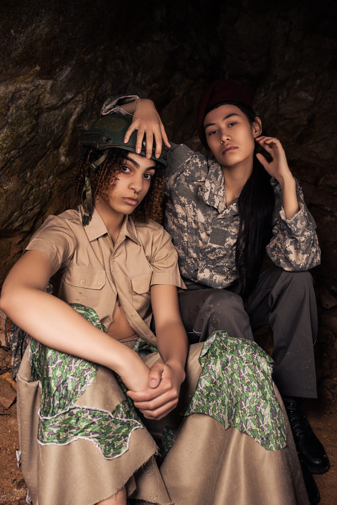 Two women in military-style clothing sitting inside a cave, one with curly hair wearing a helmet and the other with straight black hair adjusting a hat, both looking serious.