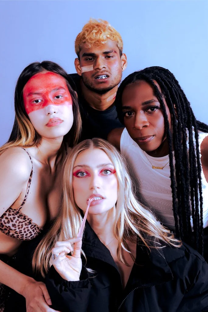 Group of four diverse young adults posing closely together, with colorful makeup and accessories, against a plain background.