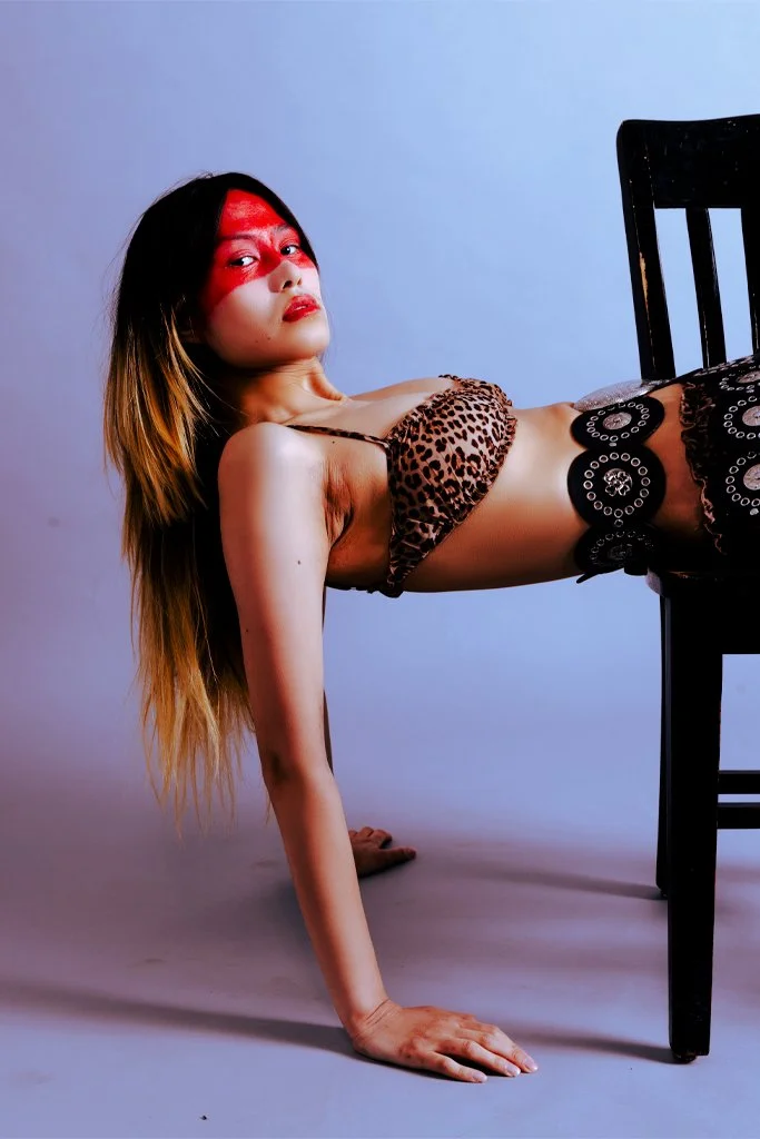 A woman with long hair and red face paint wearing leopard print clothing, posing with her hands on the ground and her legs elevated on a black chair against a plain background.