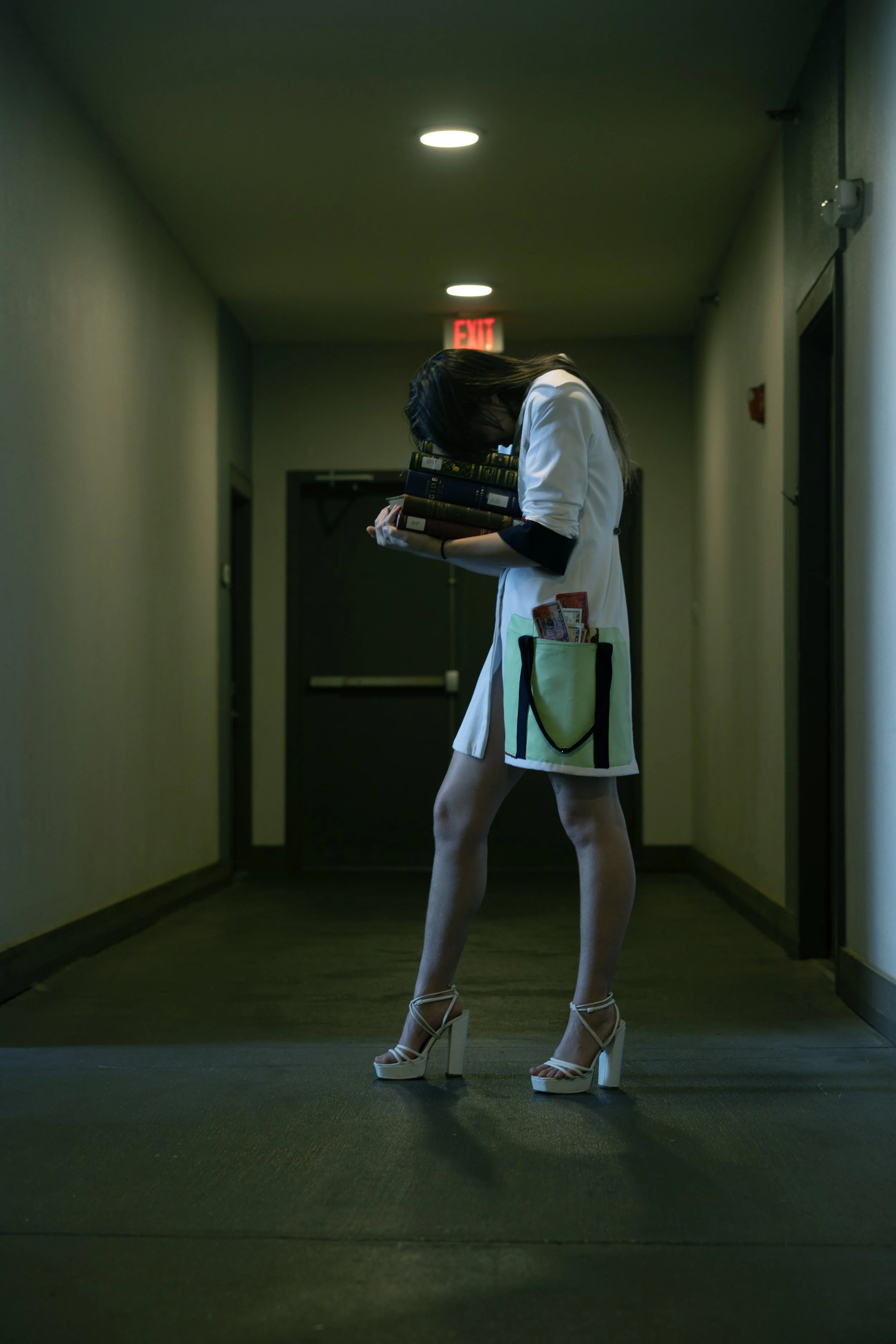 A woman in high heels and a white coat with a stethoscope pocket, appears to be a healthcare professional, standing in a hallway, holding a stack of books, with a bag containing money hanging from her waist.