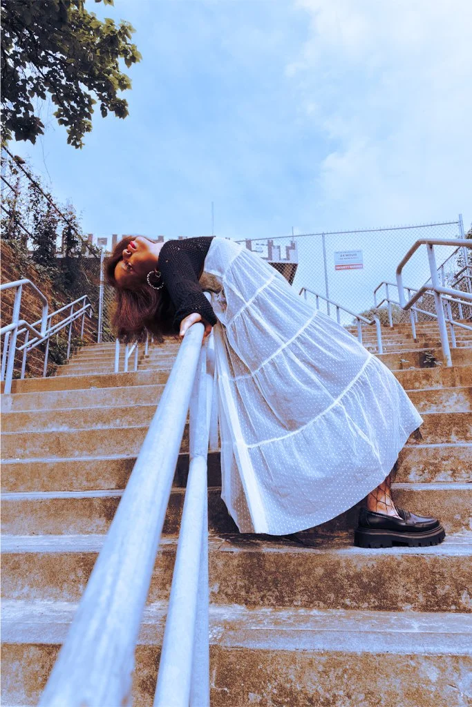 A woman leaning back on a staircase railing outdoors with sky and trees in the background, wearing a white dress, black top, and black boots.