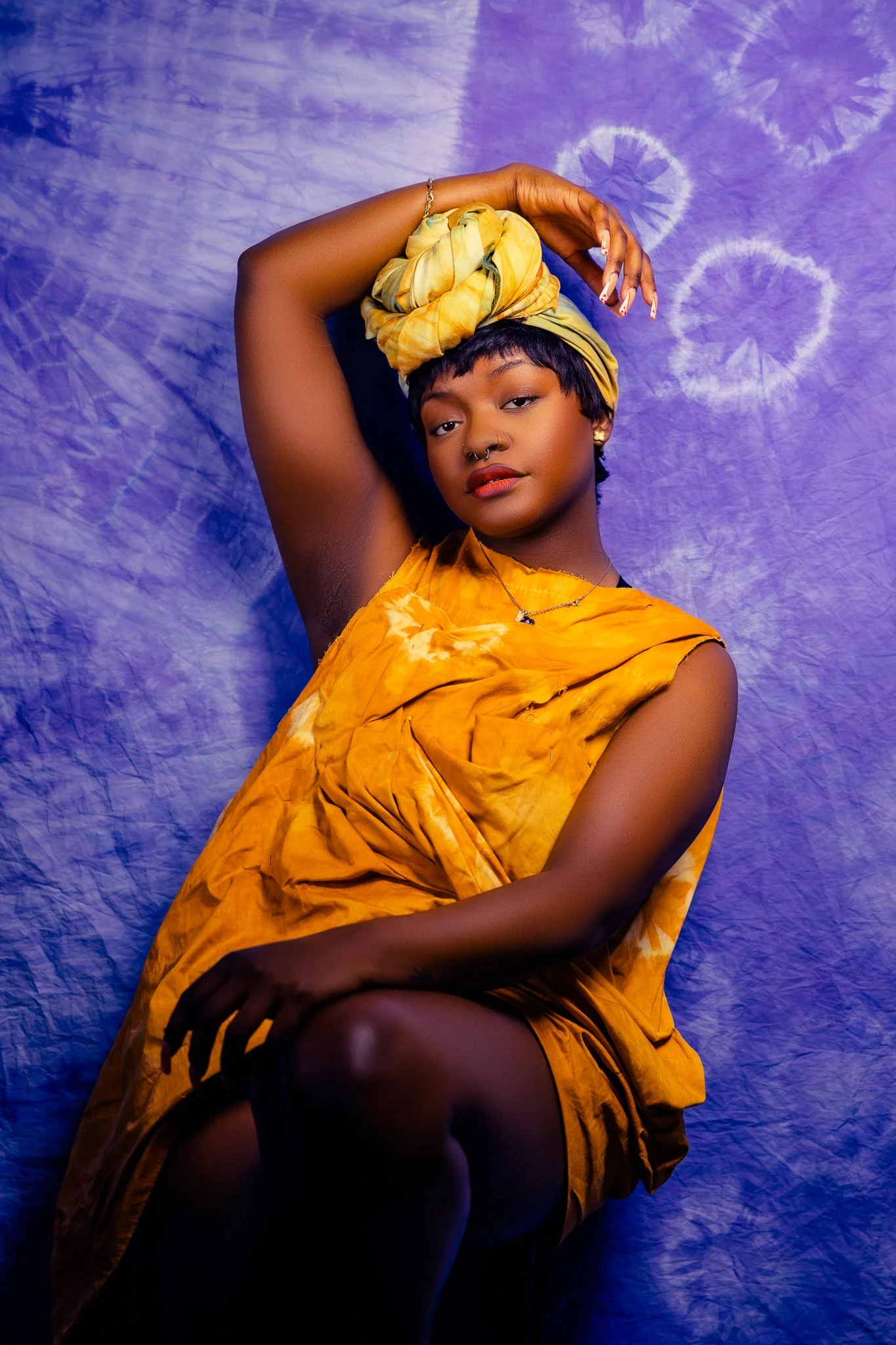 A young woman with short hair and a nose ring poses with one arm raised above her head, wearing a yellow tie-dye dress and matching headwrap, against a purple tie-dye background.