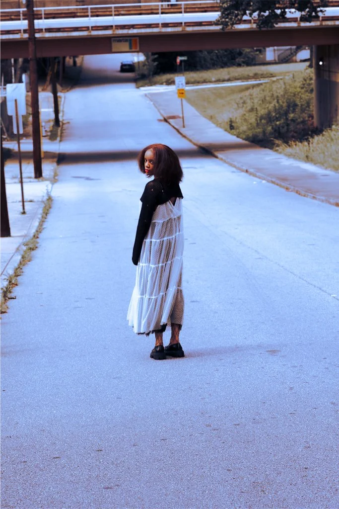 A young woman with curly hair stands on an empty street near a bridge, wearing a black top with a white sheer dress, black shoes, and looking back at the camera.