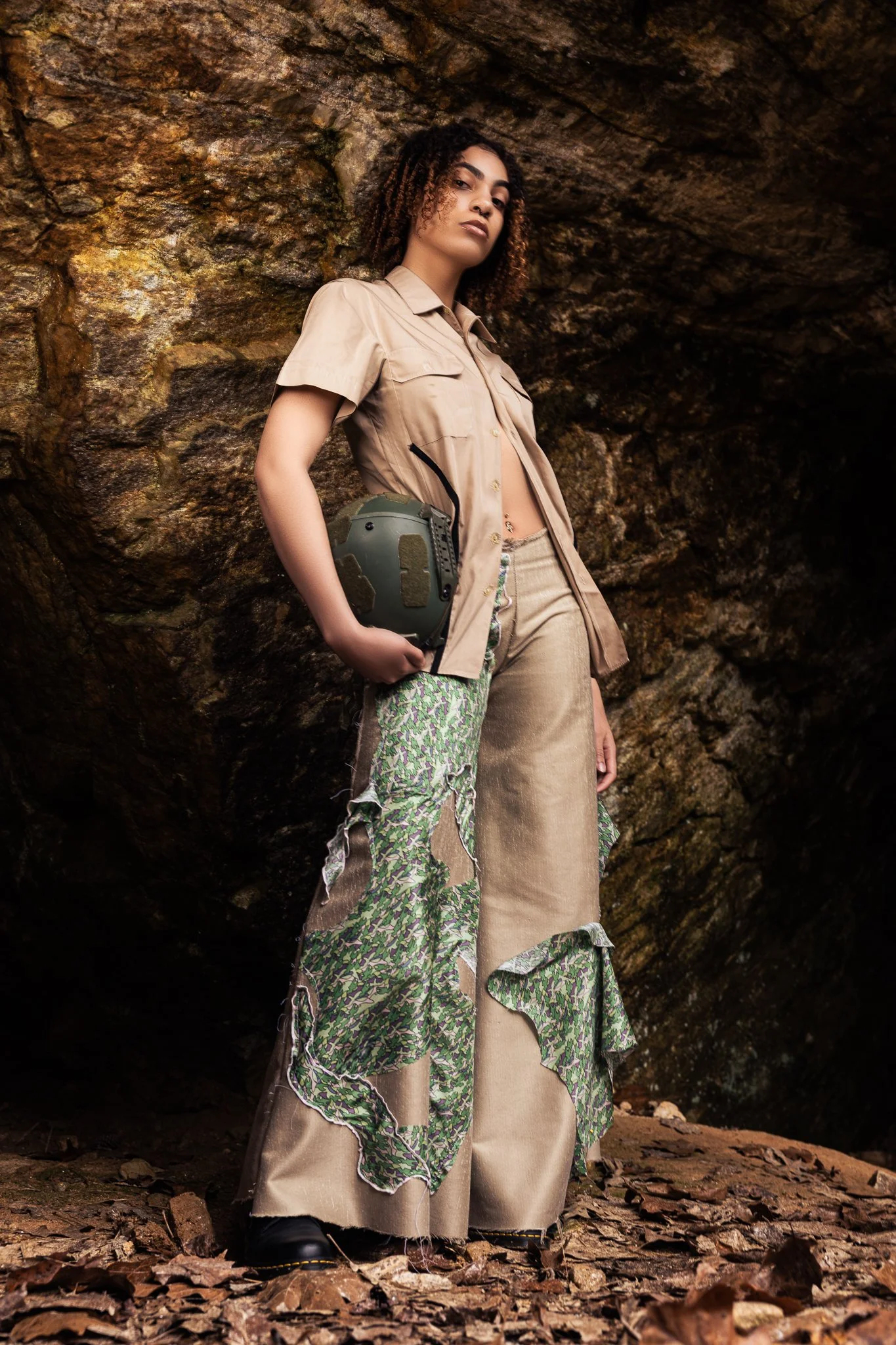 A woman holding a military helmet, standing in front of a rocky cave or opening, wearing beige shirt and wide-legged pants with green camouflage ruffles.