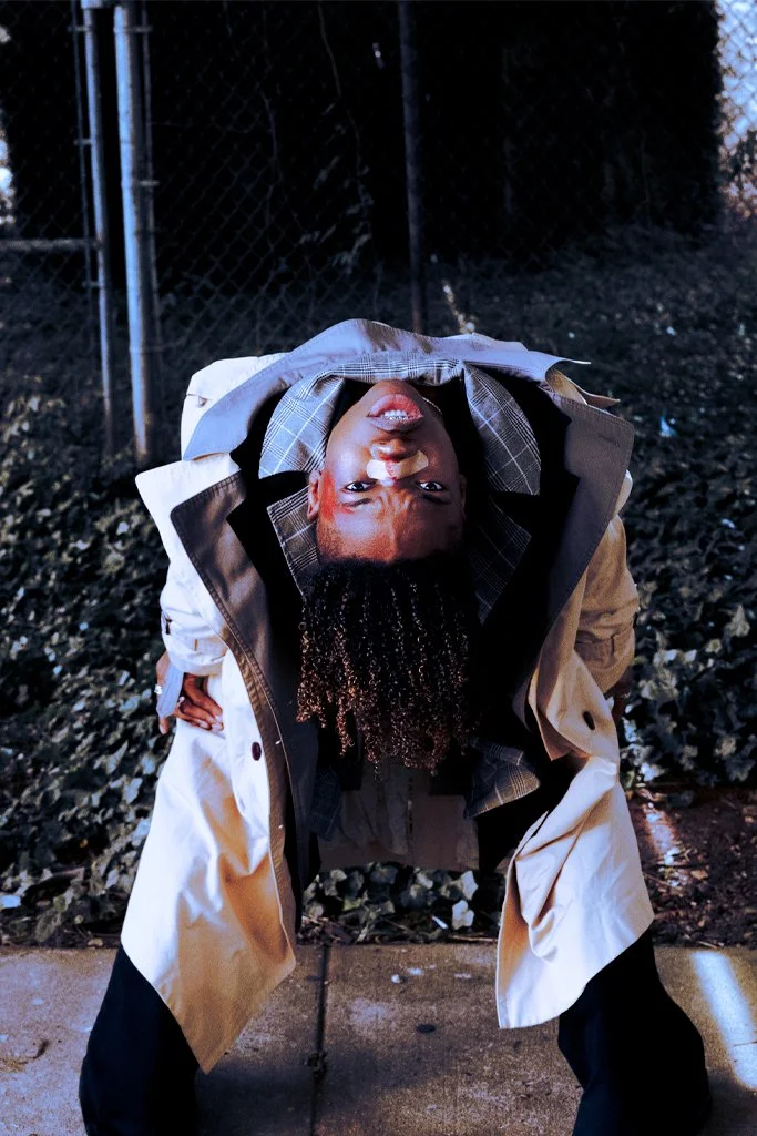 A person with curly hair bending upside down, wearing a trench coat and plaid shirt, outside at night with a chain-link fence and dark foliage in the background.