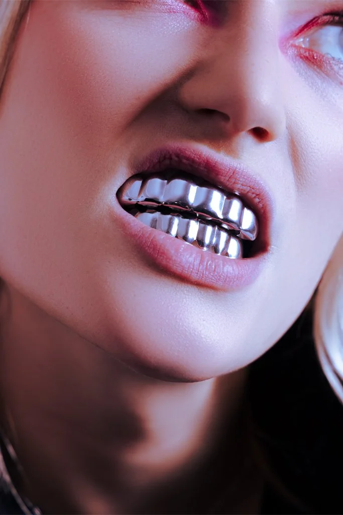 Close-up of a person's face showing metallic teeth jewelry and a slightly open mouth, with visible lips and part of the nose and eye.