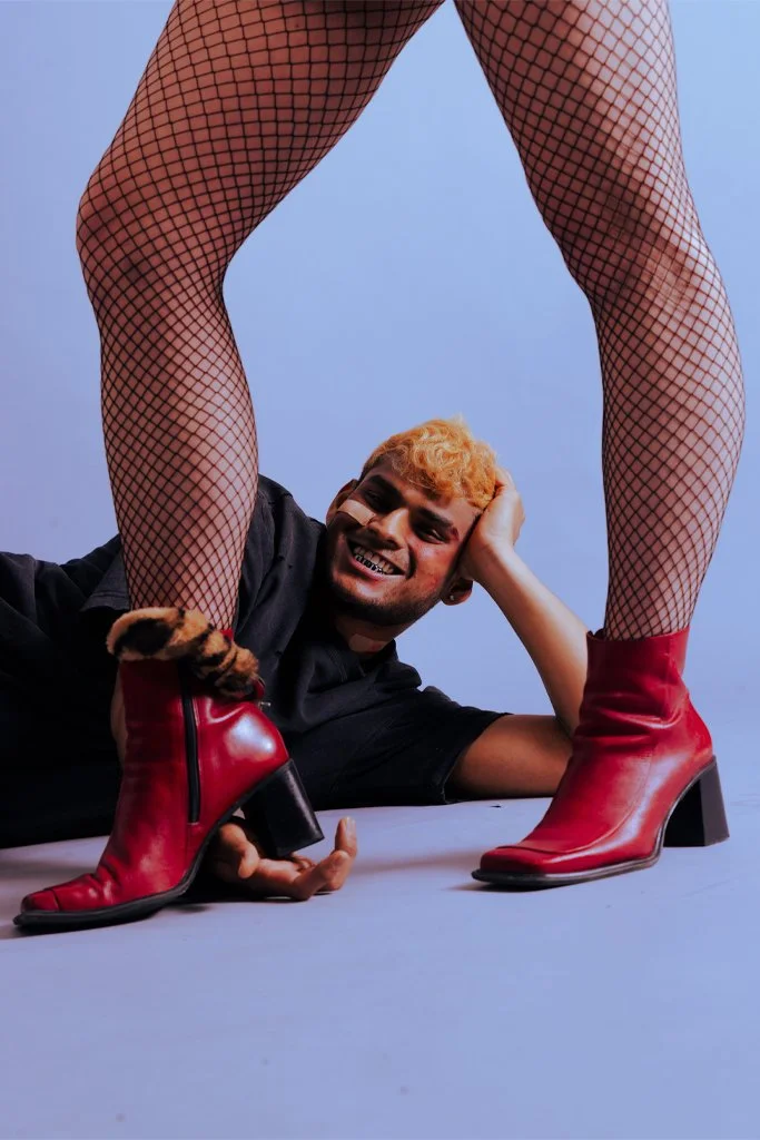A man with curly orange hair is smiling while lying on his side, supporting his head with his hand. He is positioned underneath a person wearing fishnet stockings and red boots, with the person's legs wide apart. The background is a plain, light-colo