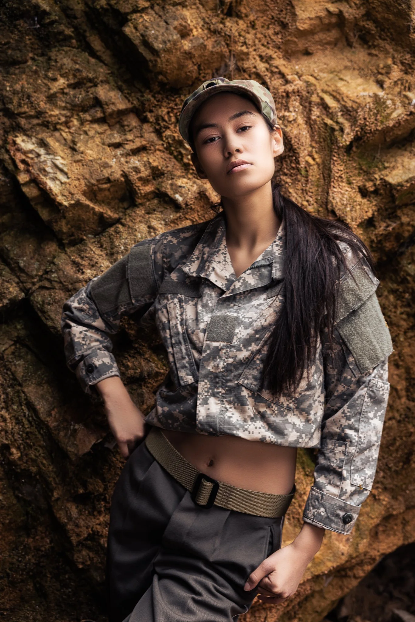 Woman in military camouflage uniform posing against rocky background.