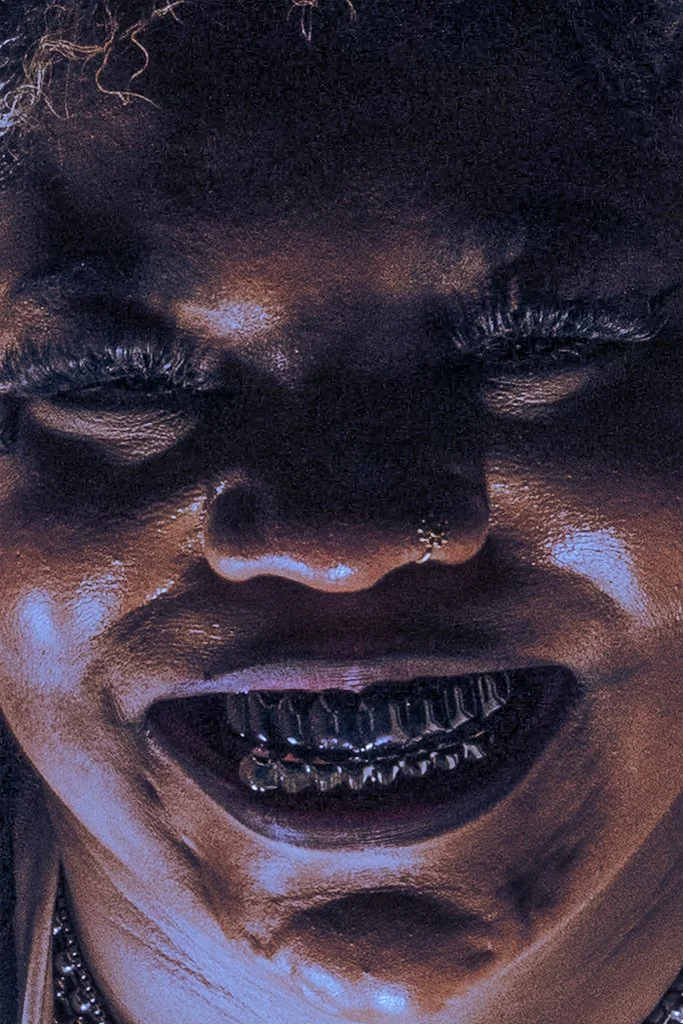 Close-up of a person with dark skin, smiling with visible teeth and wearing a grill, with their eyes partially closed.