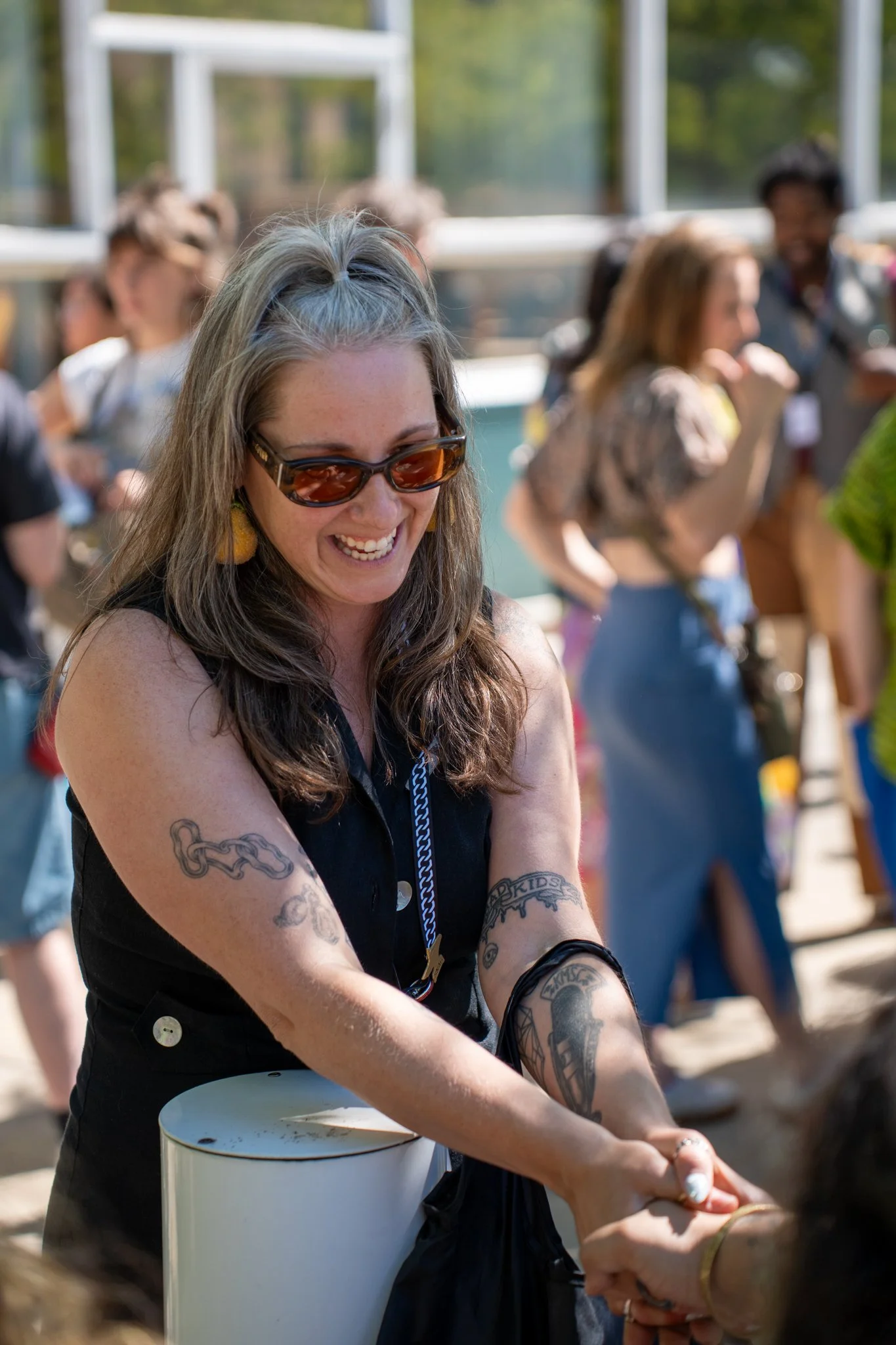 A woman with long gray hair, wearing sunglasses and tattoos on her arms, is smiling and playing a drum outdoors at a gathering or festival, with several people blurred in the background.