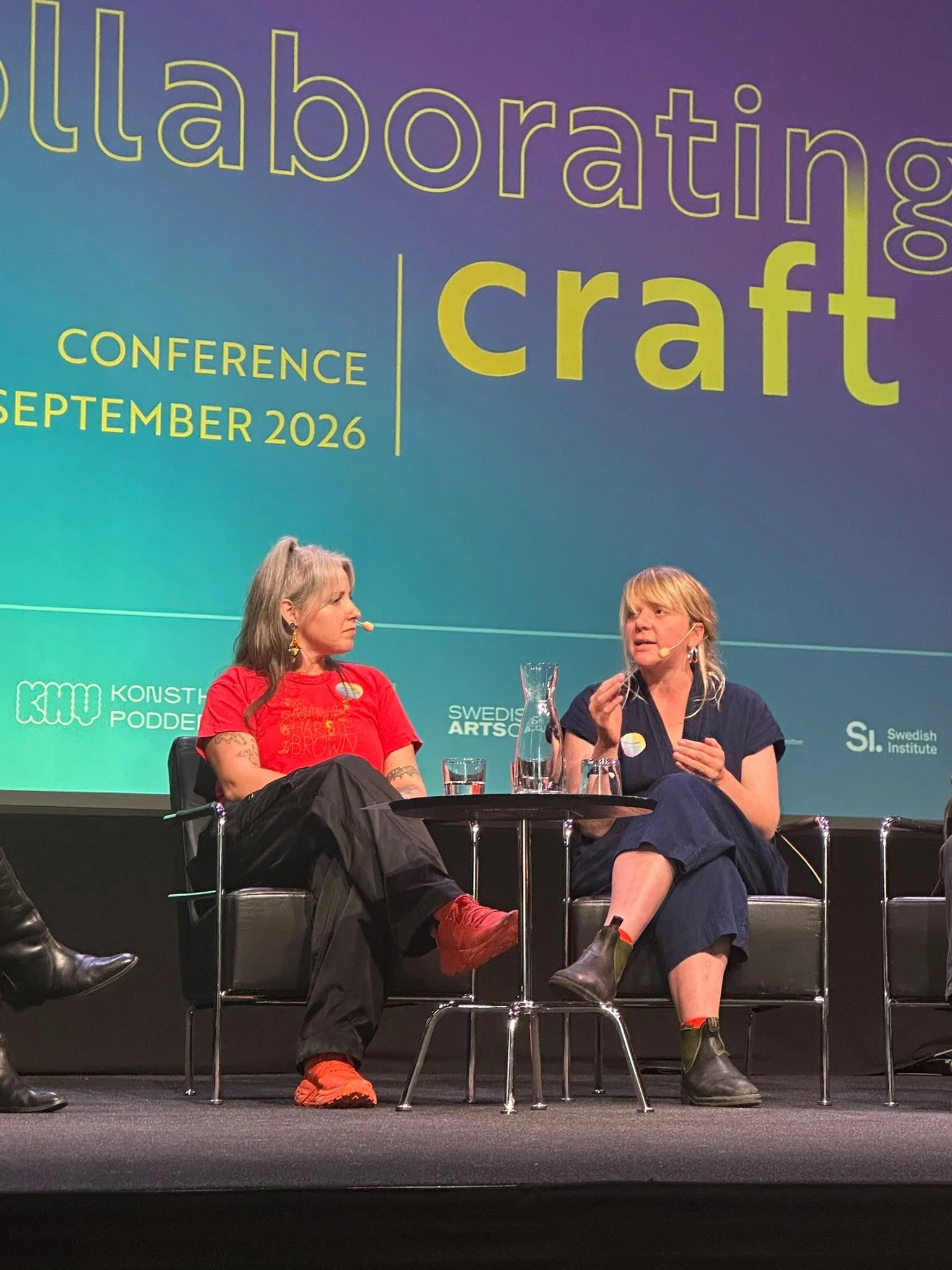 Two women seated on stage at a conference, engaged in conversation, with a large presentation screen behind them displaying text about an event happening in September 2026.
