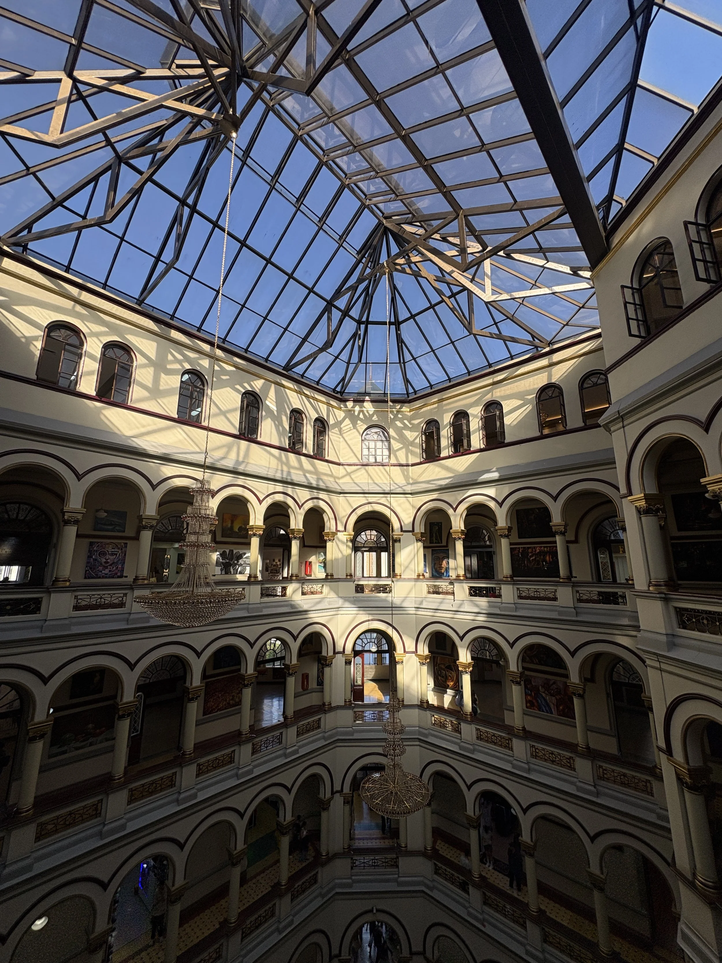 Ornate multi-story atrium with arched galleries, chandeliers, artwork, and a glass-paned skylight.
