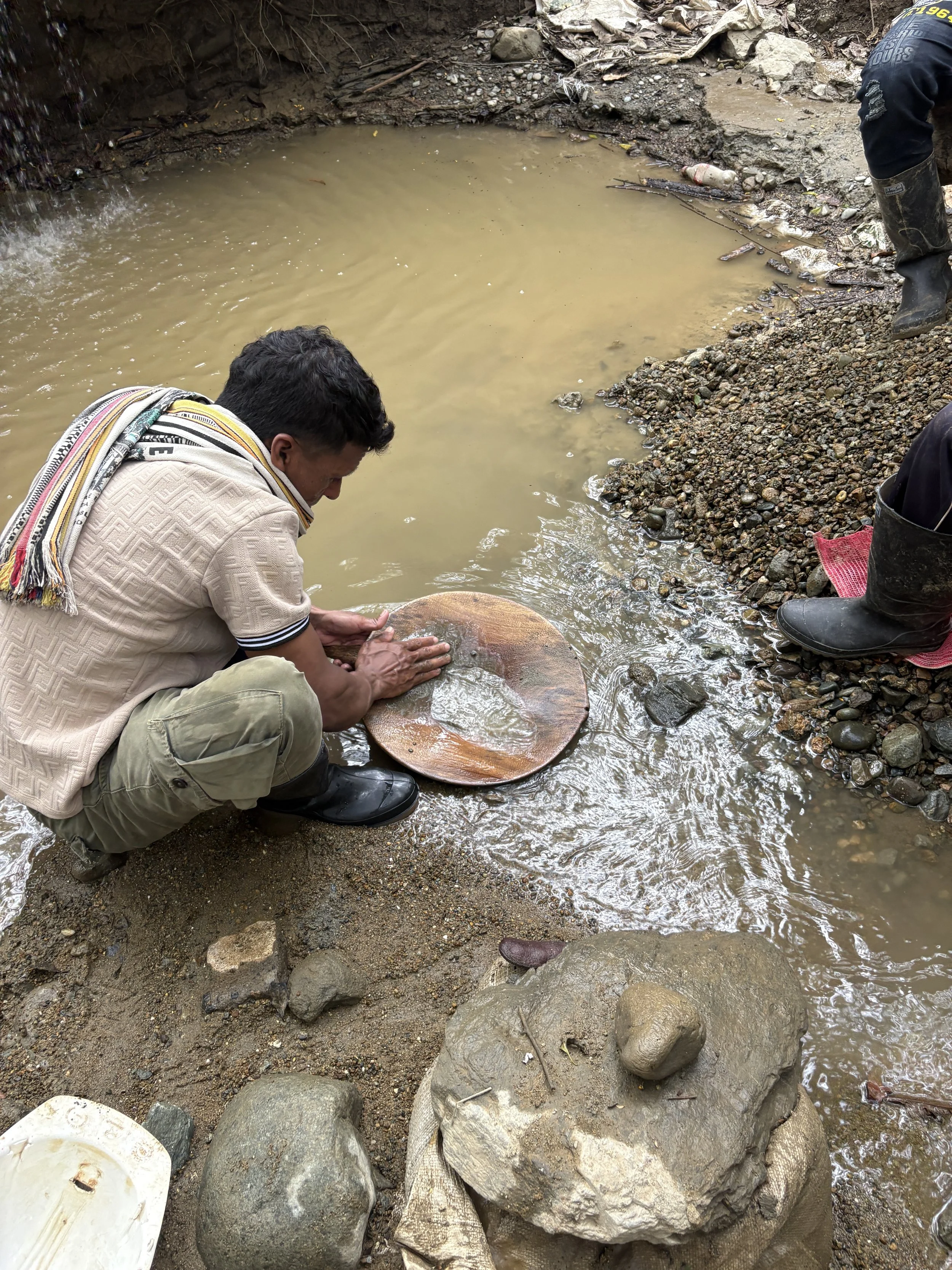 People wash a large wooden gold-panning tray in a muddy creek beside a small waterfall.