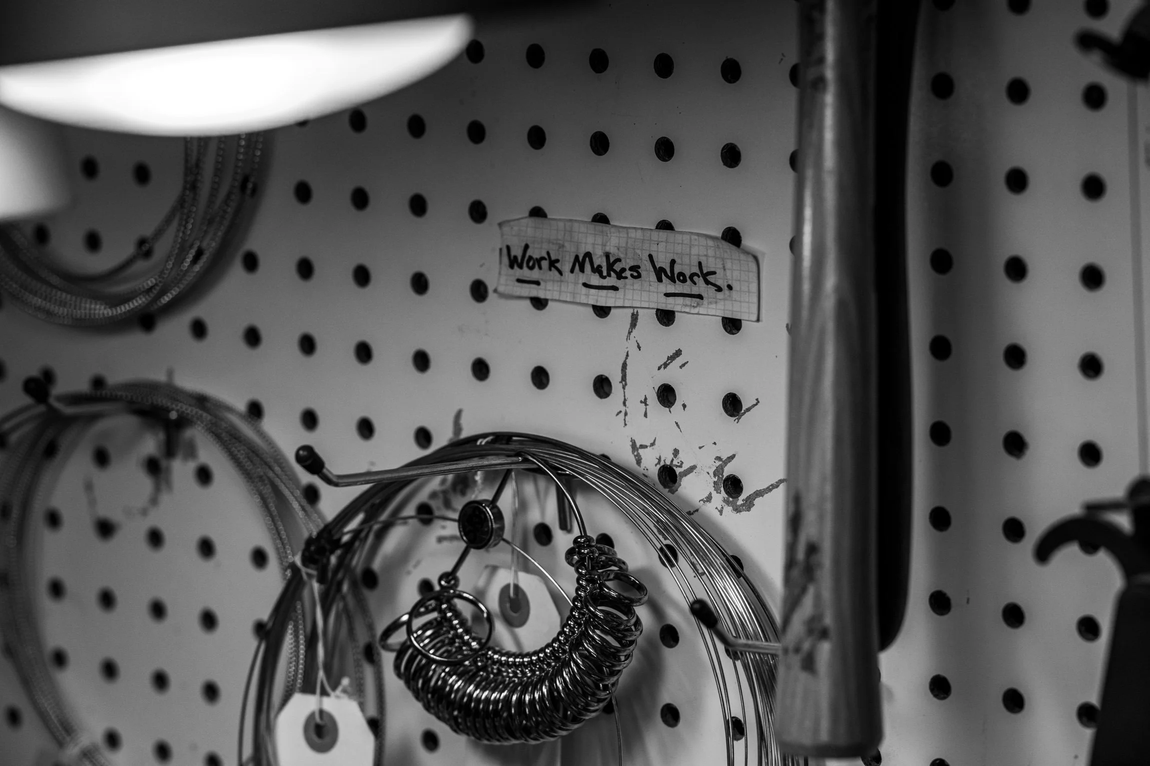 Black and white photo of Sarah Rachel Brown’s jewelry workbench, featuring coiled metal wire, tools, and a handwritten note reading “Work Makes Work,” symbolizing her journey from artist to storyteller and digital marketer.