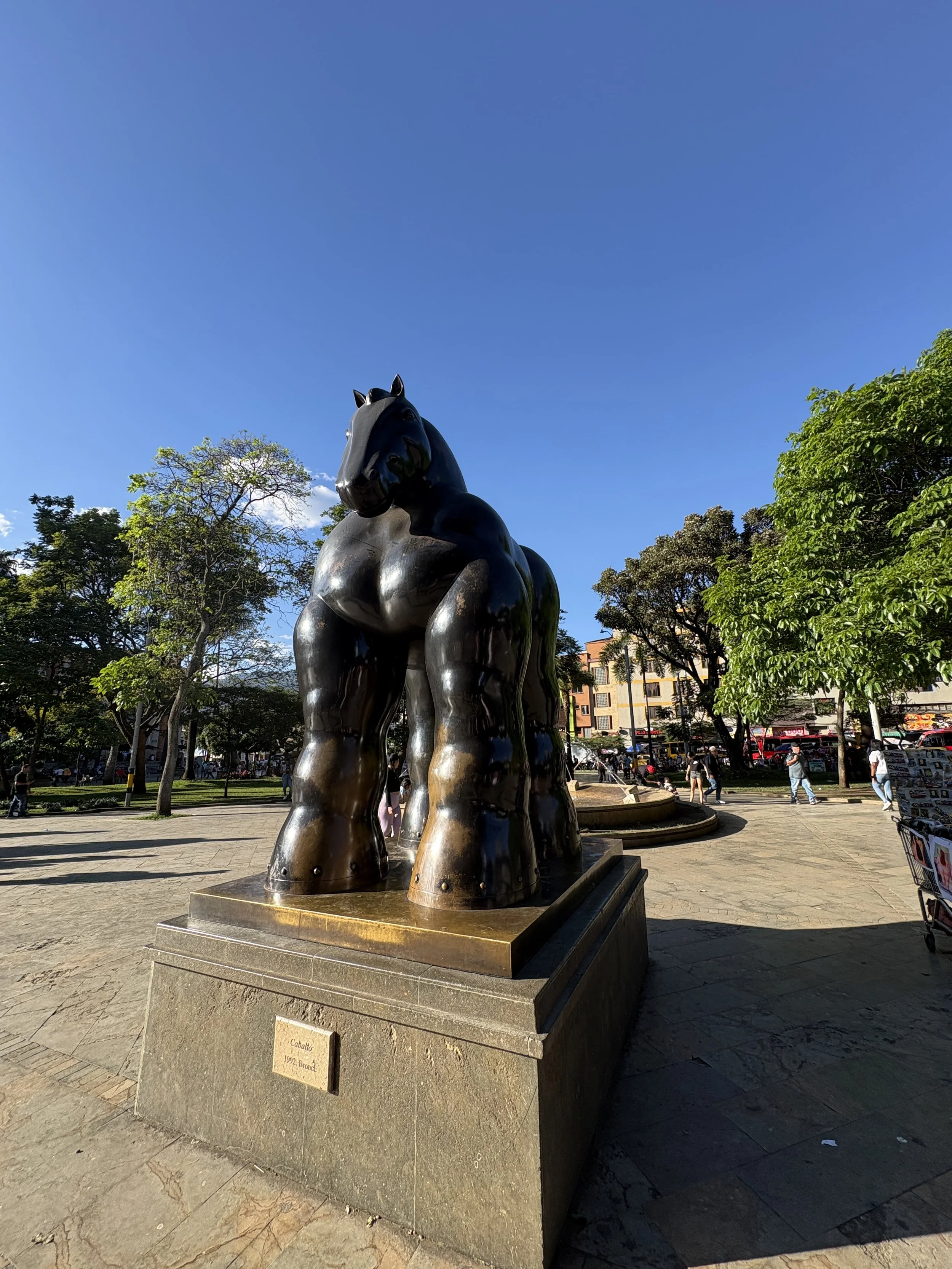 Bronze horse sculpture by Fernando Botero in a tree-lined city plaza under a clear blue sky