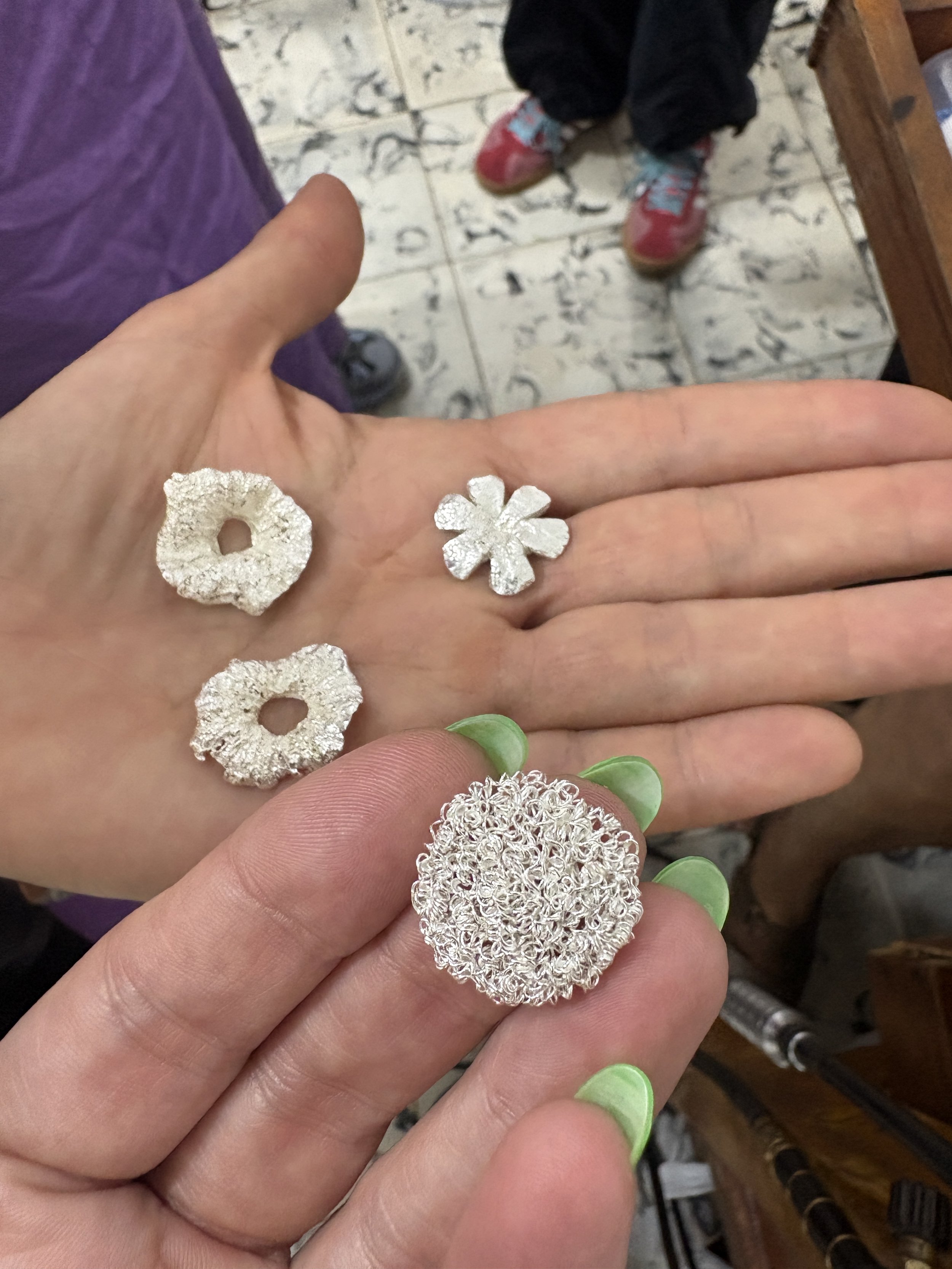 Close-up of a hand displaying textured silver jewelry pieces, including flower-shaped and circular ornaments, with green-painted fingernails.