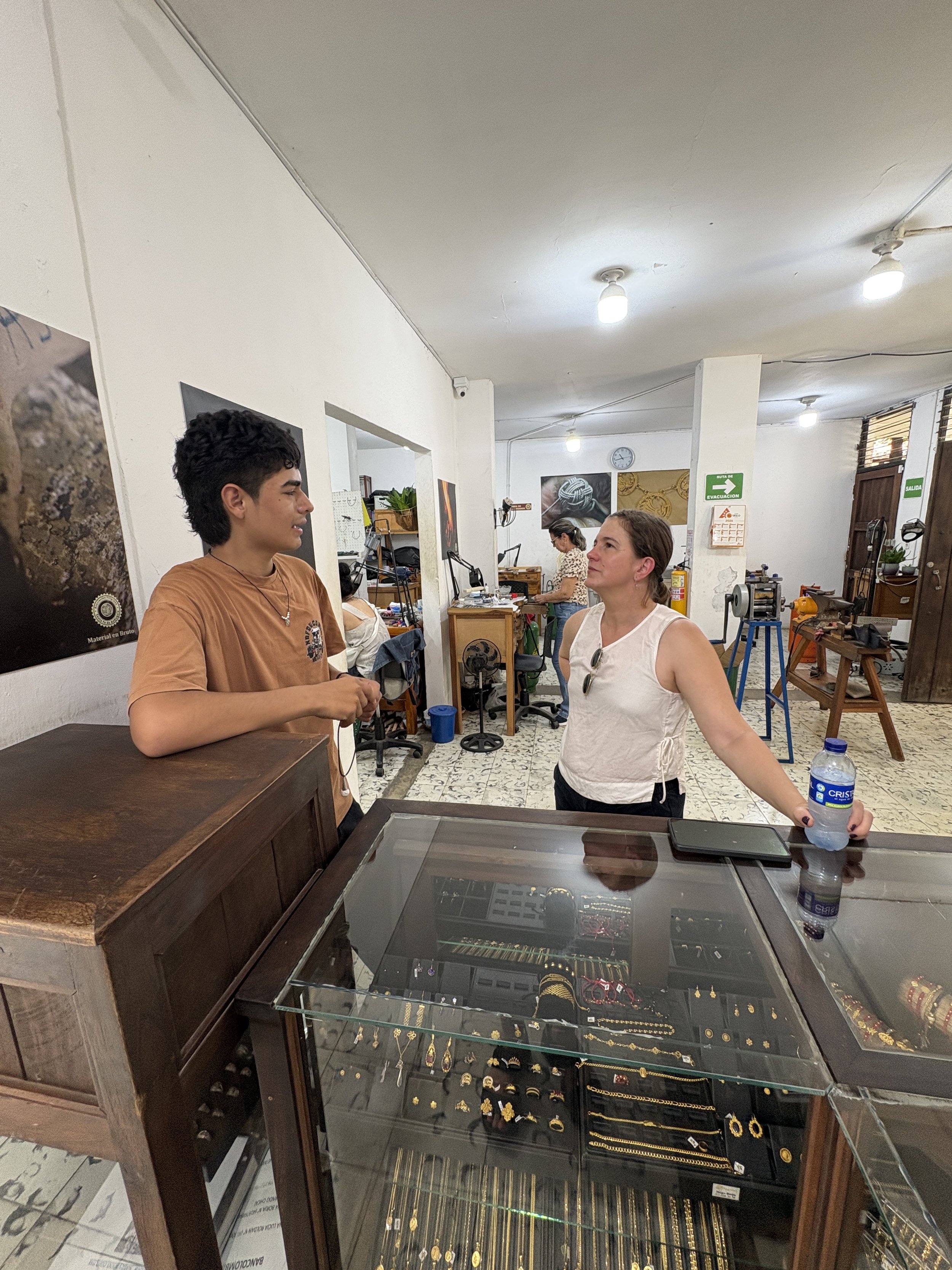 People inside a jewelry workshop or store, with glass display cases filled with gold jewelry, tools, artwork, and ceiling lights.