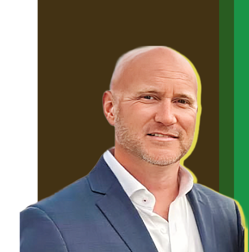 A bald man with a beard wearing a navy suit jacket and white shirt, smiling against a brown background with green accents.