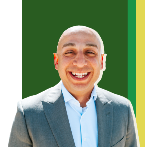 A bald man smiling and wearing a light blue dress shirt and gray suit jacket, standing in front of a green background with yellow and green vertical stripes on the right side.