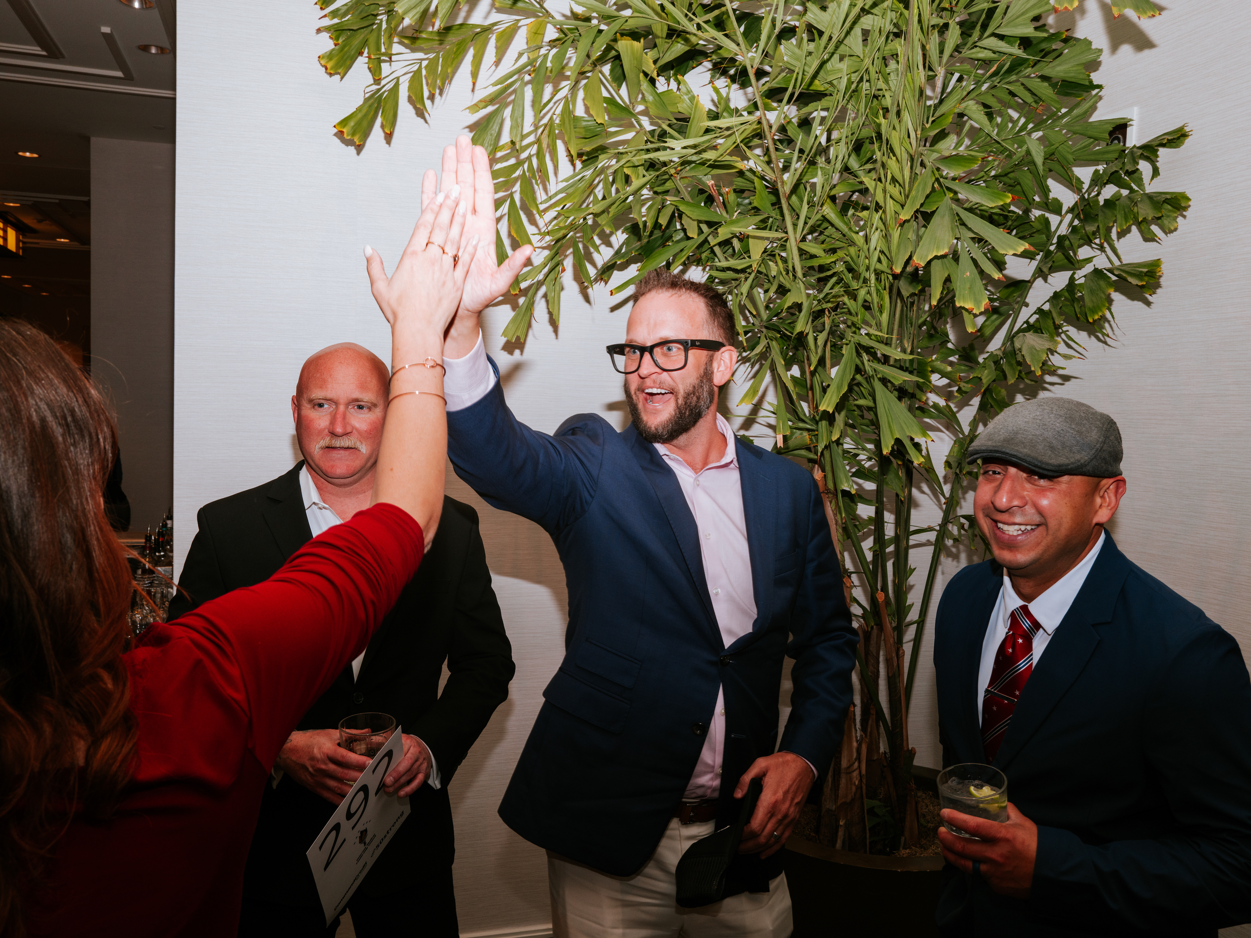 Four VA Loan Nerd team members at a social event, high-fiving and smiling, standing near a large potted plant.
