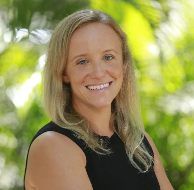 A woman with long blonde hair and blue eyes, smiling, wearing a black sleeveless top, outdoors with greenery background.