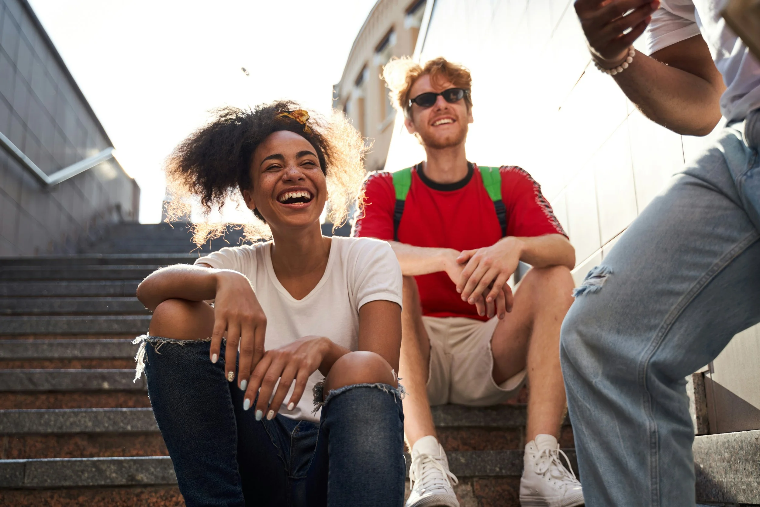 Three young people sitting on outdoor stairs, laughing and smiling, with buildings in the background.