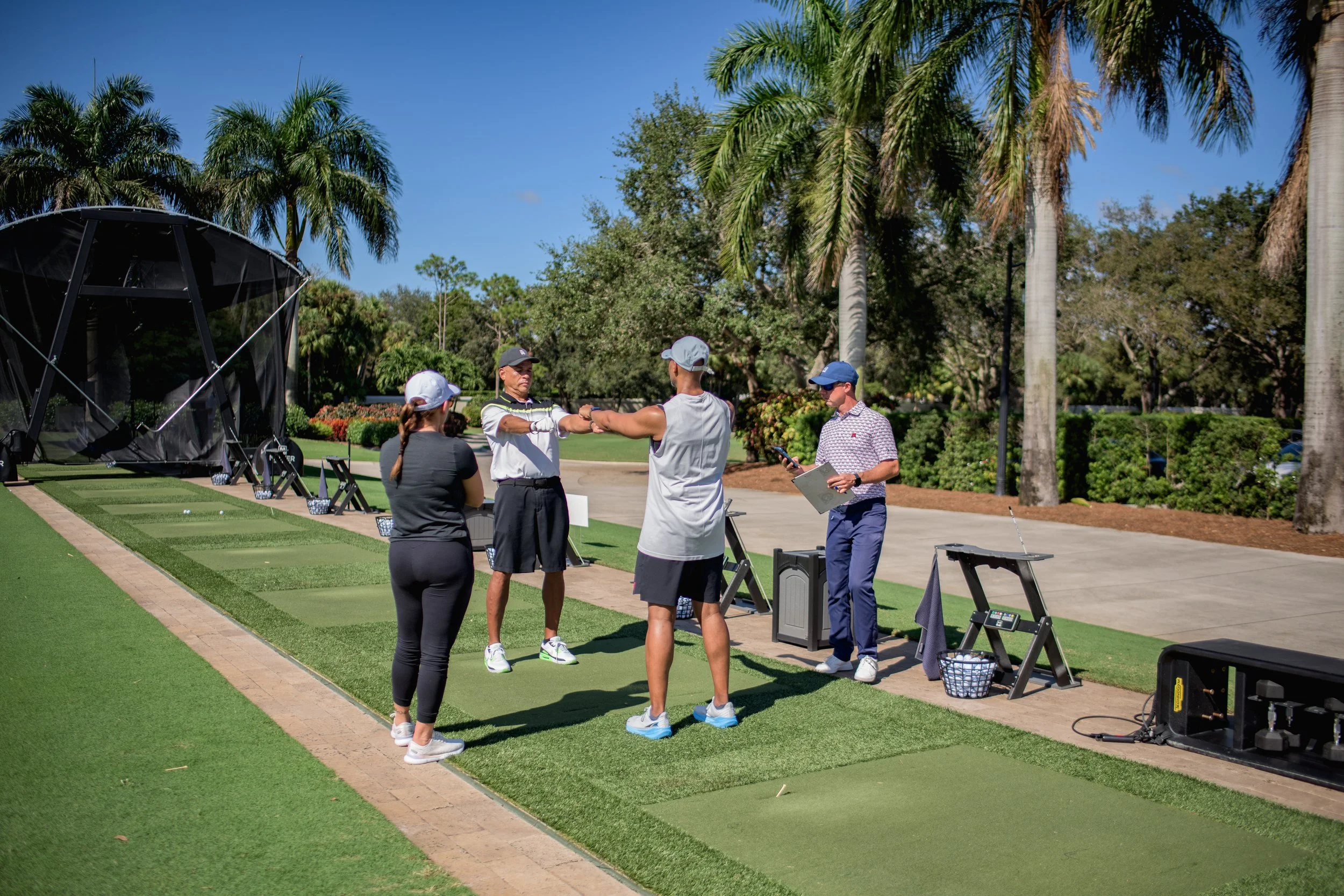 A golf instructor demonstrating a swing to three students at a driving range with palm trees and clear blue sky in the background.