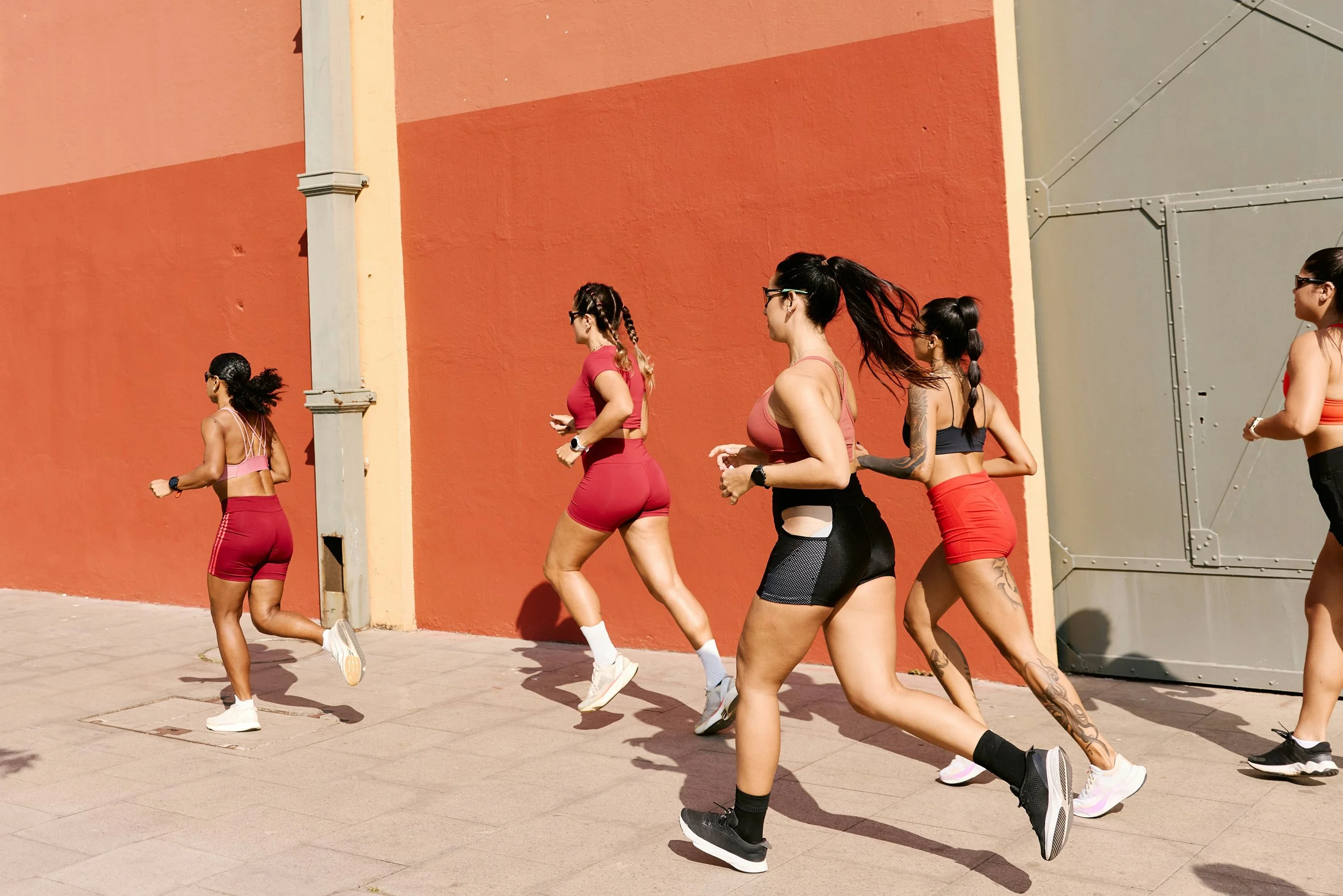 Six women jogging outdoors on a sunny day, dressed in athletic wear, with a large orange and gray building wall in the background.