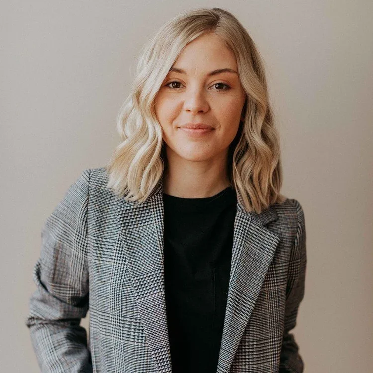 A young woman with blonde wavy hair wearing a gray plaid blazer and a black top, standing against a light-colored wall and smiling softly.