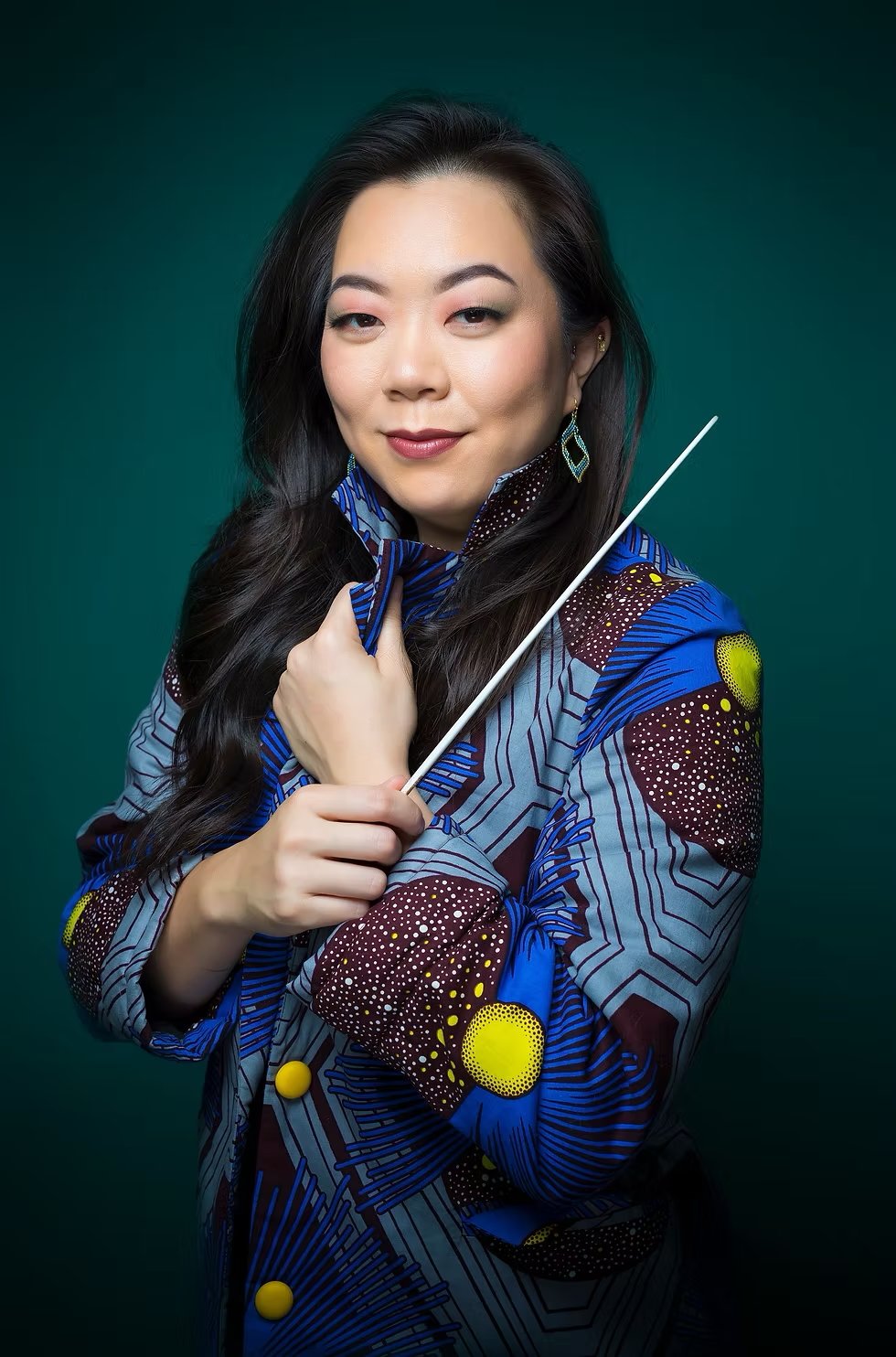 Portrait of a woman with long black hair, wearing a colorful patterned jacket, holding a conductor's baton, against a dark green background.