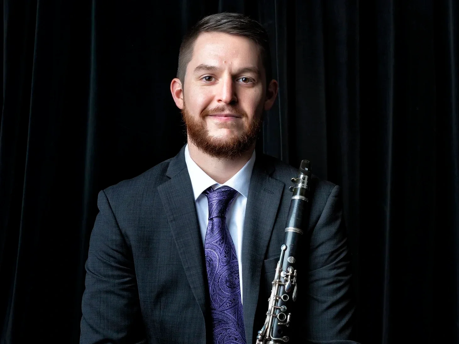 A young man in a suit holding a clarinet, standing in front of a black curtain.