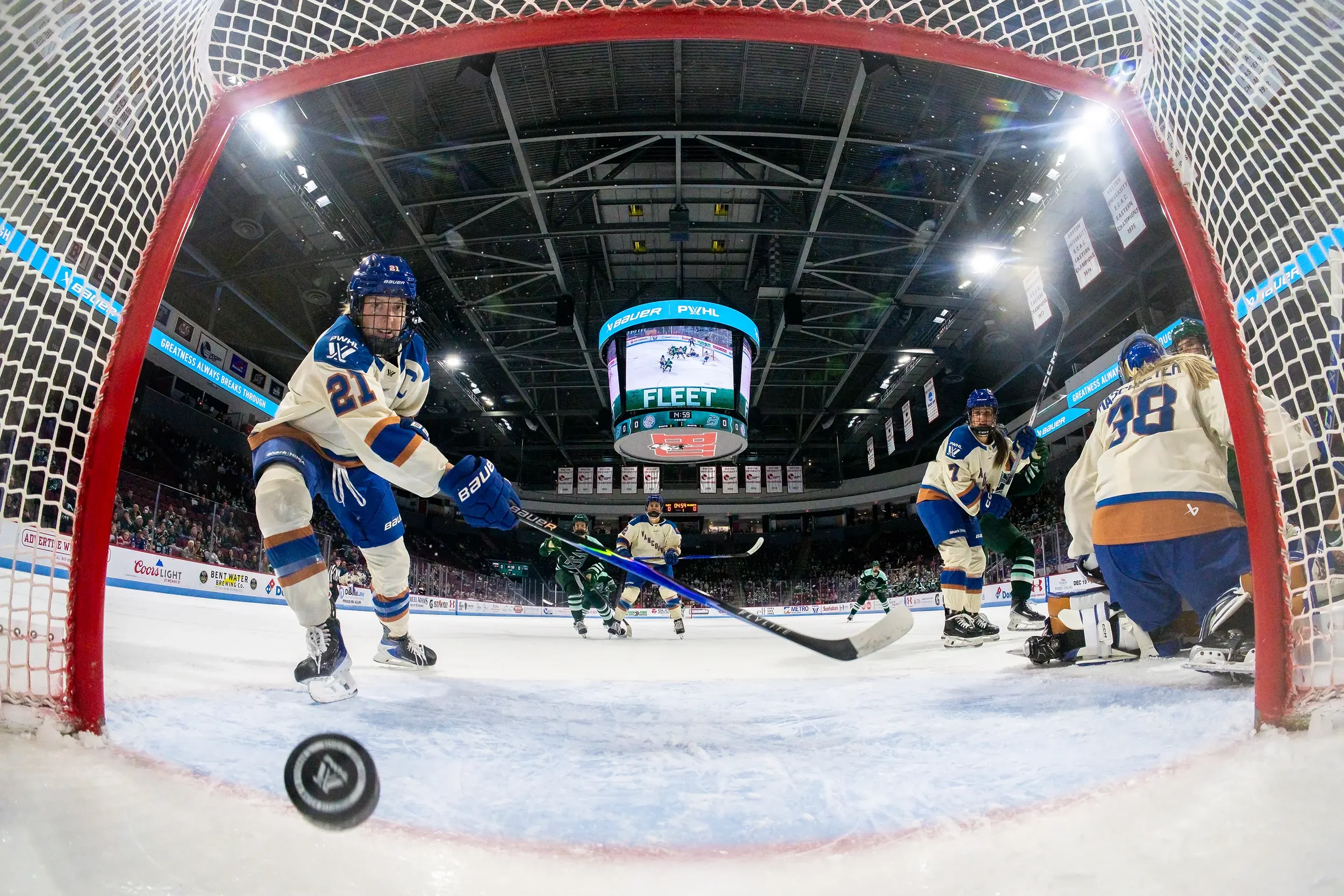 Hockey players on ice rink during game, viewed from behind the goal, with puck near the goal line, scoreboard in background.