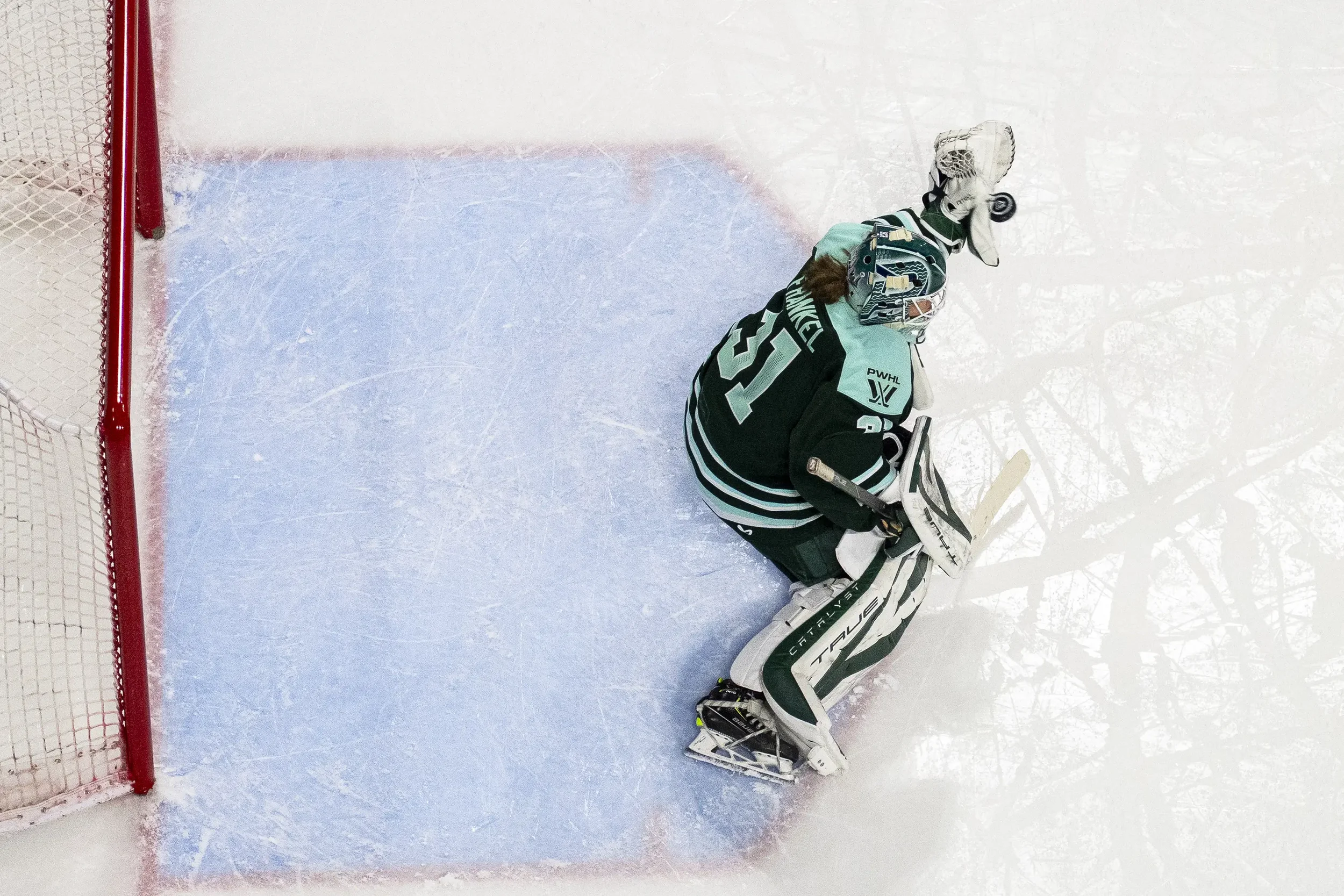 Hockey goalie kneeling in front of the net on the ice rink, wearing a teal and black uniform, protective gear, and a helmet.