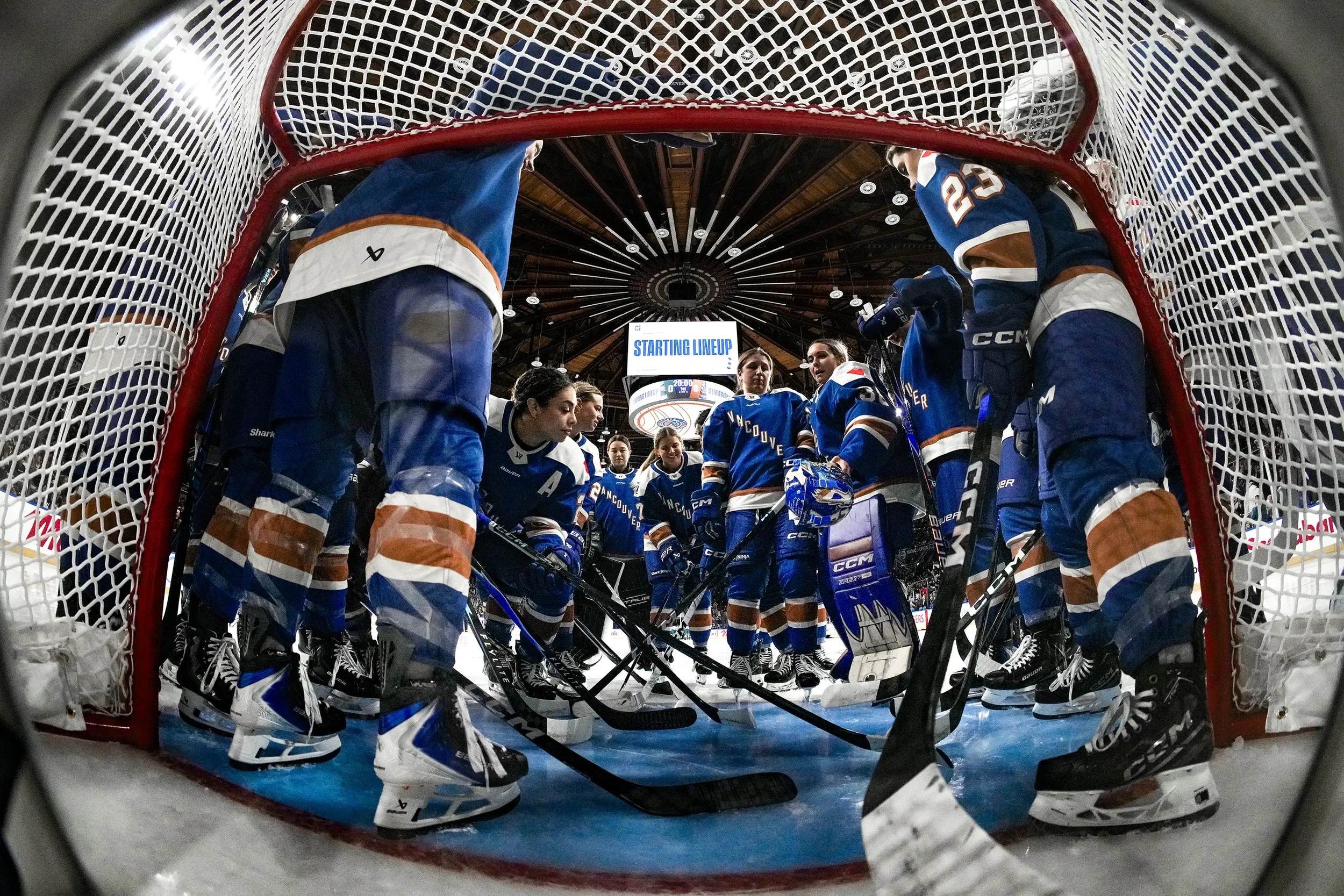 Hockey team in a huddle on the ice, viewed from inside the goal net at the rink.