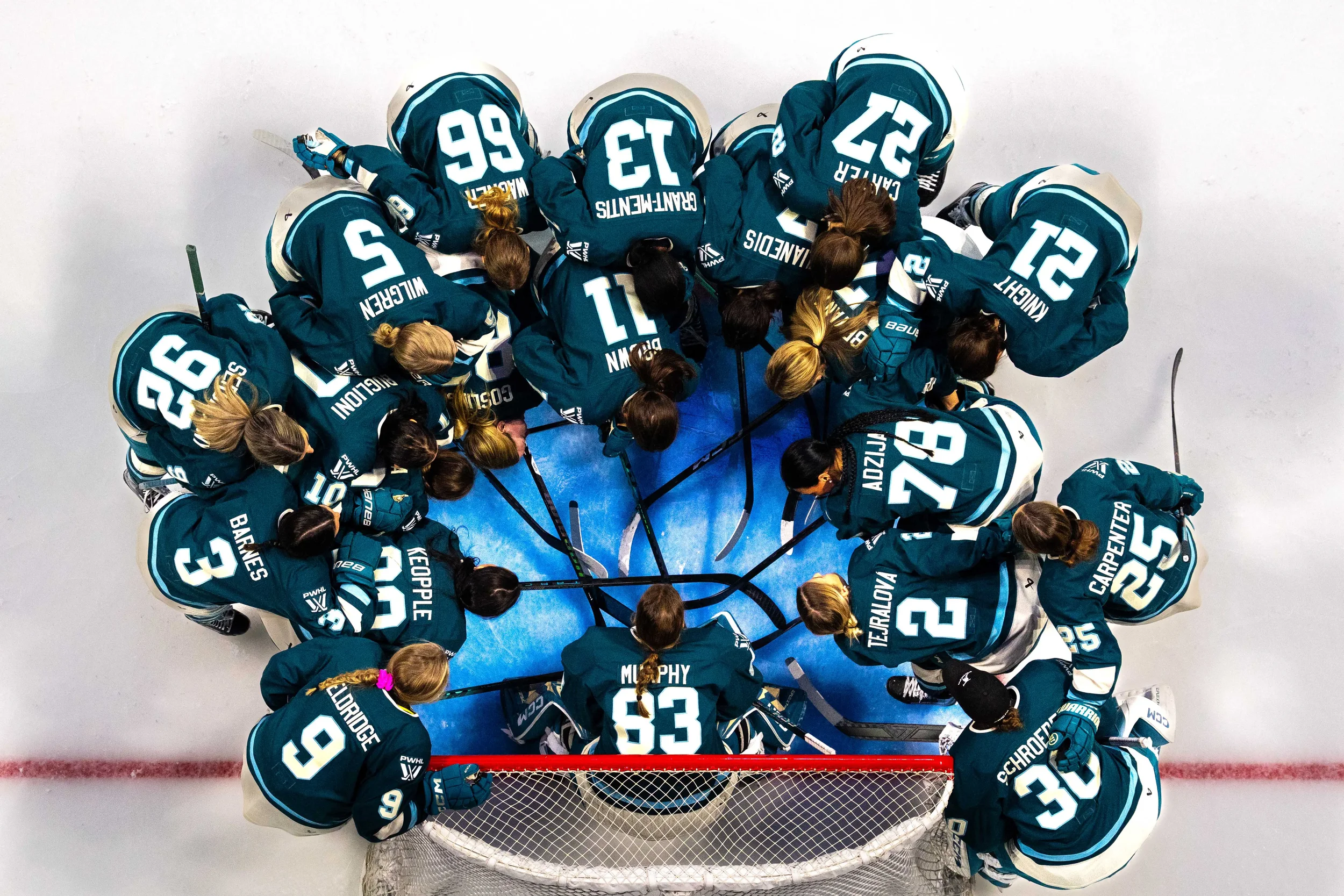 An ice hockey team wearing teal jerseys with white and black accents, gathered in a circle around the goal, with their heads bowed and hockey sticks pointing inward, on an ice rink.