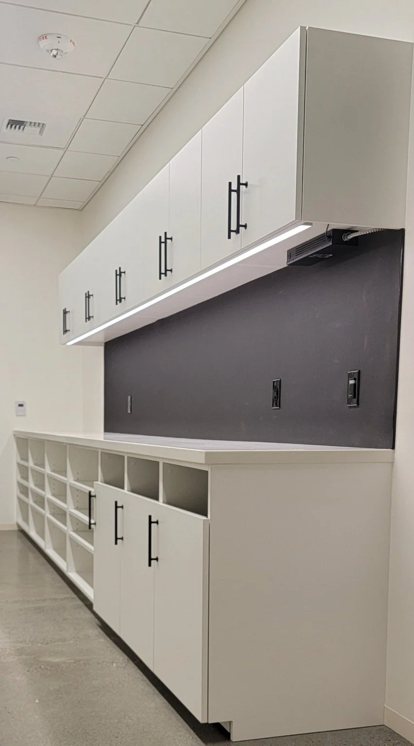 Modern cabinetry and shelving unit in a room, with white upper cabinets, black handles, and a white lower cabinet with open cubbies, a black wall with electrical outlets, and a few mounted devices on the ceiling.