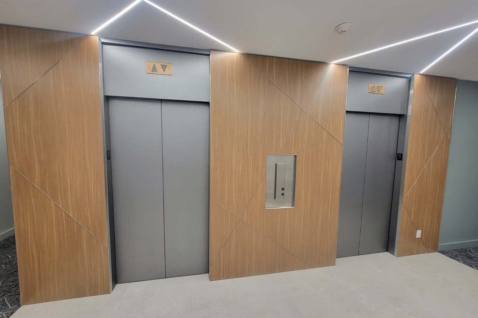 Two gray elevator doors in a modern wood-paneled wall with minimal lighting overhead and a small metal service panel between them.