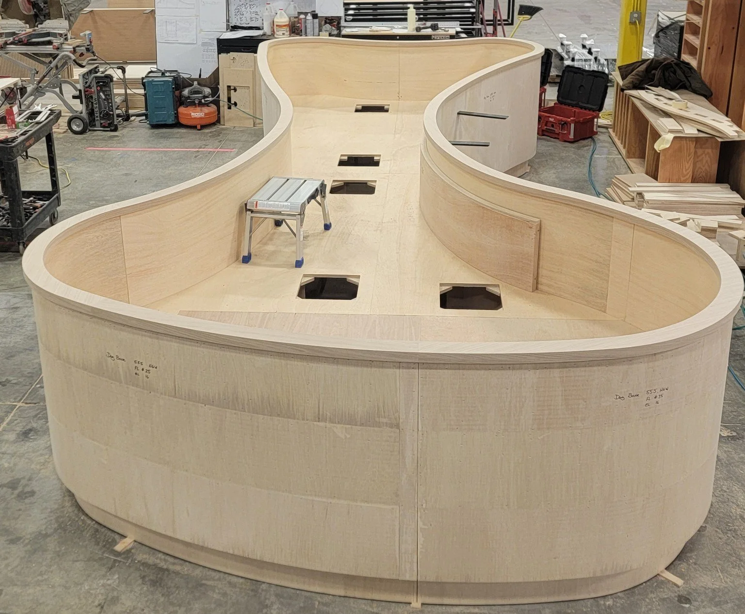 A large, curved wooden structure under construction in a workshop, with cutouts in the top surface and walls on the sides.