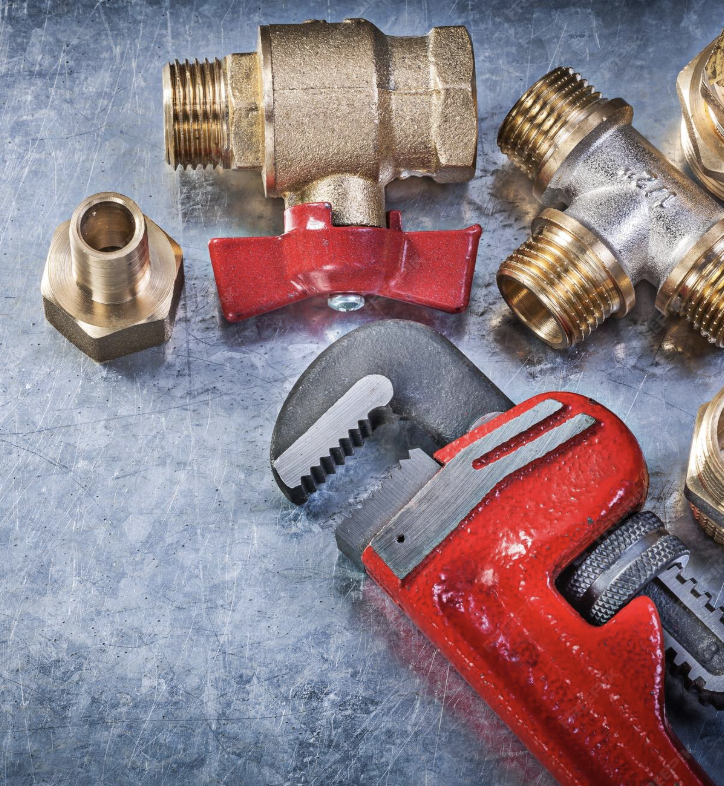 A red pipe wrench, a brass valve, and various brass fittings are placed on a metallic work surface.