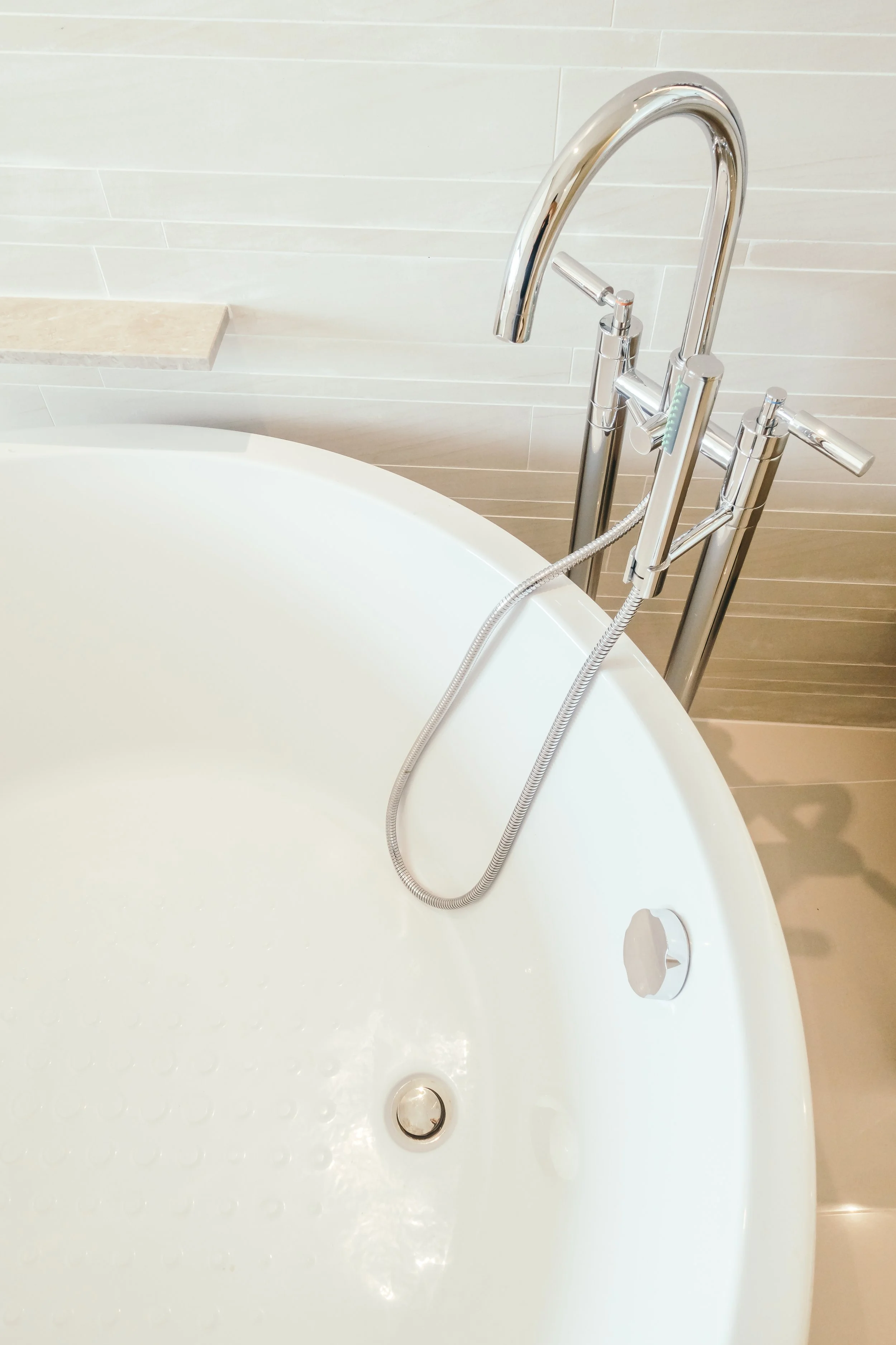Close-up view of a modern bathtub with a chrome handheld showerhead attached to a metal stand, a faucet, and a circular drain, with beige tiled wall in the background.