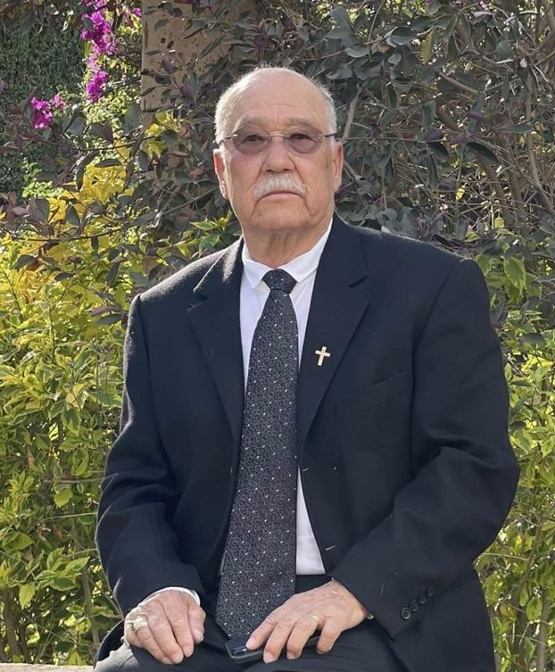 An elderly man with glasses and a serious expression, dressed in a black suit, white shirt, and dark tie with white specks, sitting outdoors in front of a bush with green leaves and purple flowers.