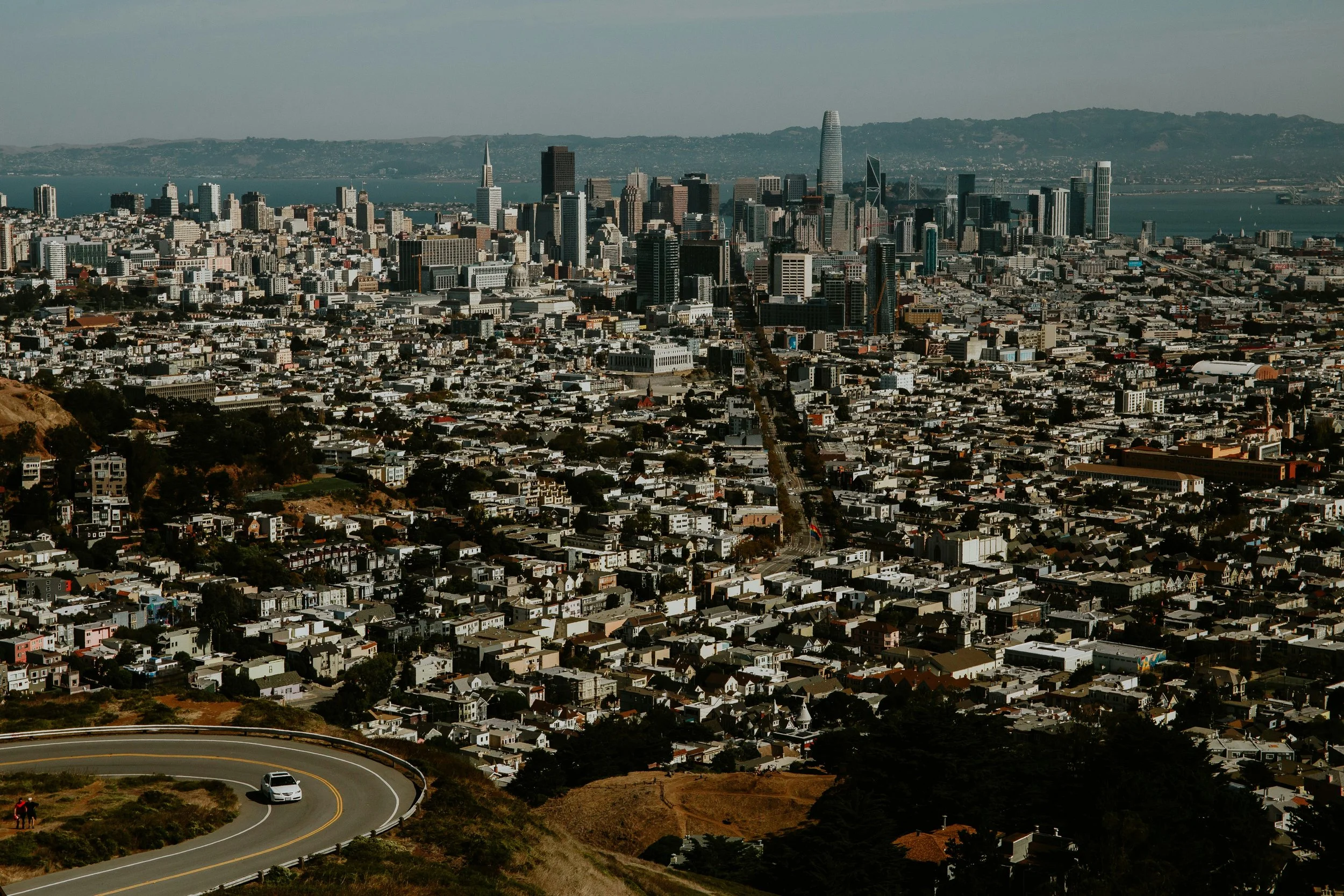 Aerial view of San Francisco cityscape with downtown skyscrapers, winding streets, and a scenic hilly area in foreground, near the Water, under a partly cloudy sky.