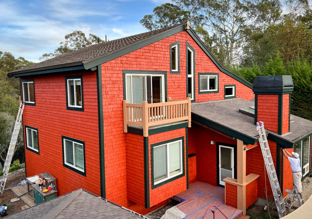 A large, multi-story house painted red with black trim, surrounded by trees. The house has various windows, a balcony, and a sloped roof. Two workers are on ladders working on the house exterior.