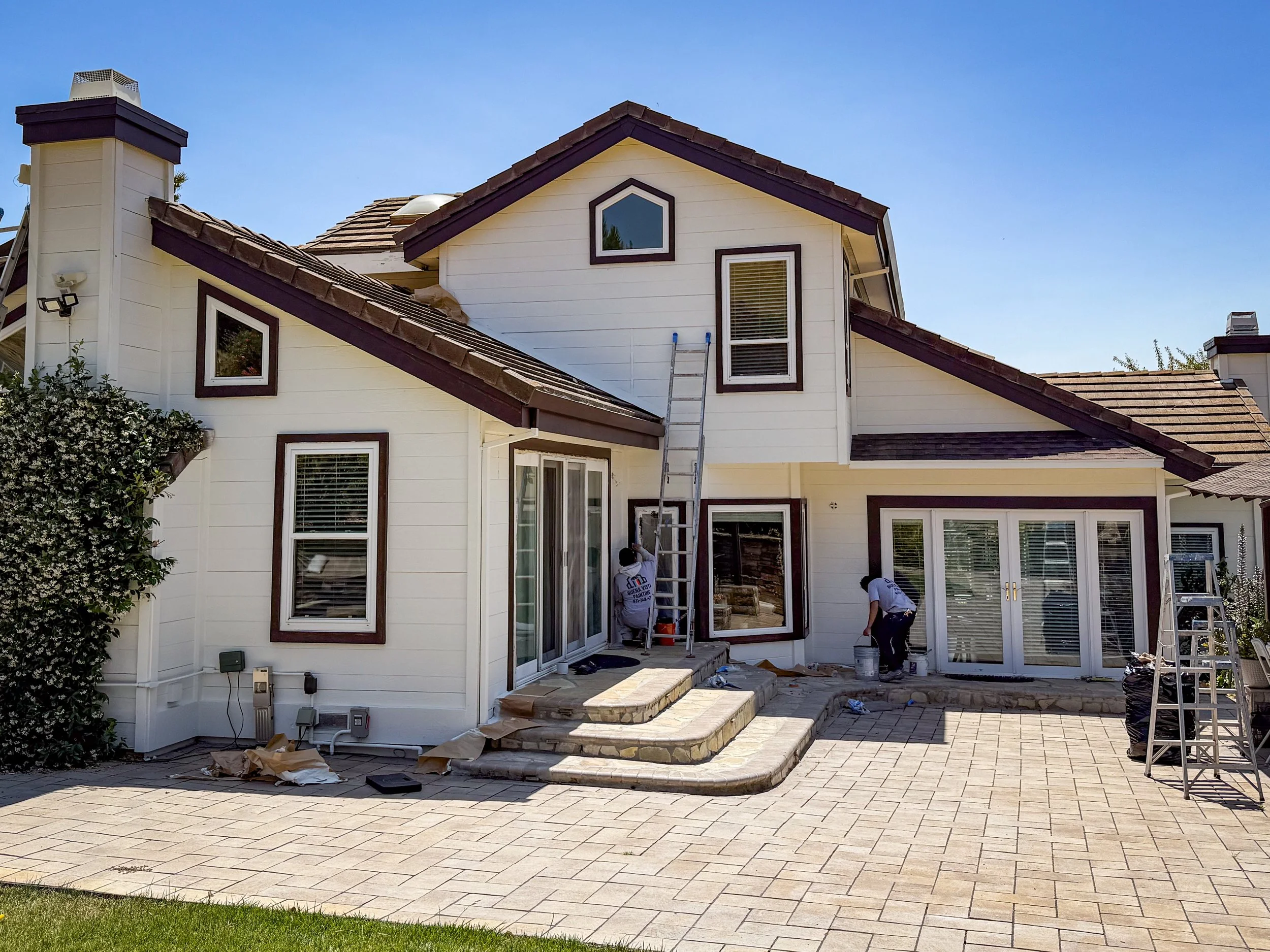 A white two-story house with brown trim and a shake tile roof under construction or renovation. Workers are seen working on the house’s exterior, with ladders and construction materials around. There is a paved patio area in the foreground.